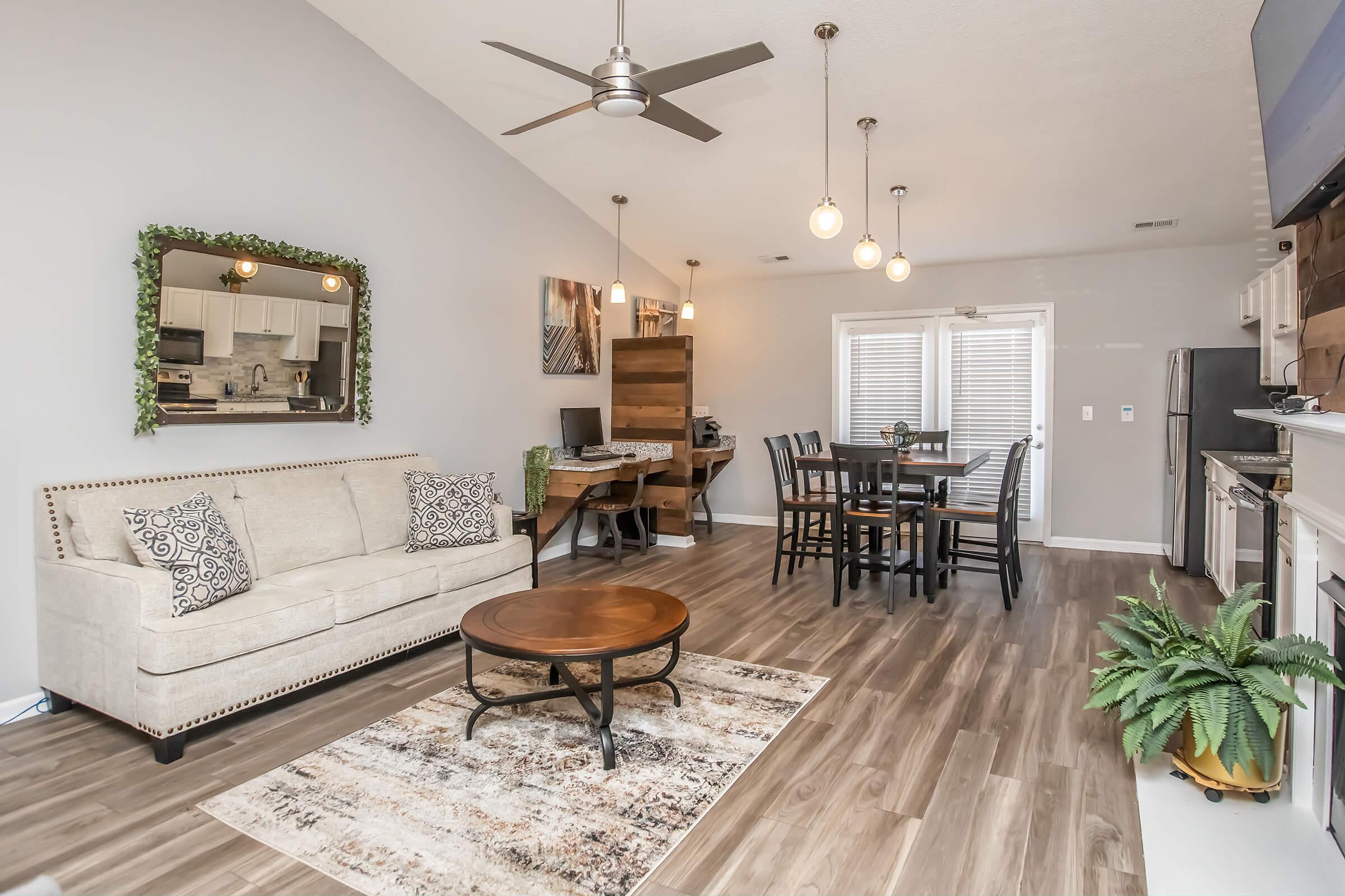 A modern, open-concept living area featuring a beige sofa with decorative pillows, a round coffee table, and a stylish rug. The space includes a dining table with black chairs, a ceiling fan, and large windows allowing natural light. Greenery adds a touch of warmth to the contemporary decor.