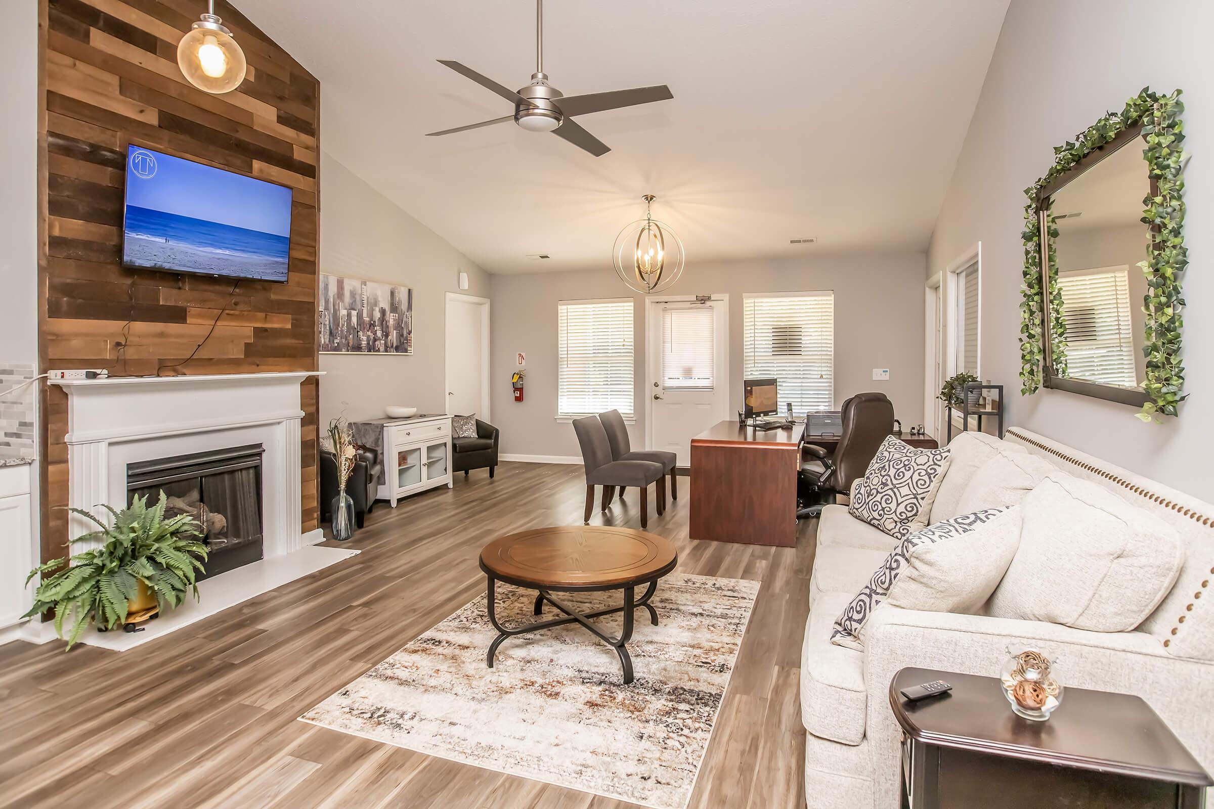 Living room featuring modern decor with a beige sofa, round wooden coffee table, and a large flat-screen TV mounted on a wooden accent wall. There are two chairs, a desk area, and a fireplace. Natural light enters through windows, and there are plants and a mirror adding to the cozy ambiance.