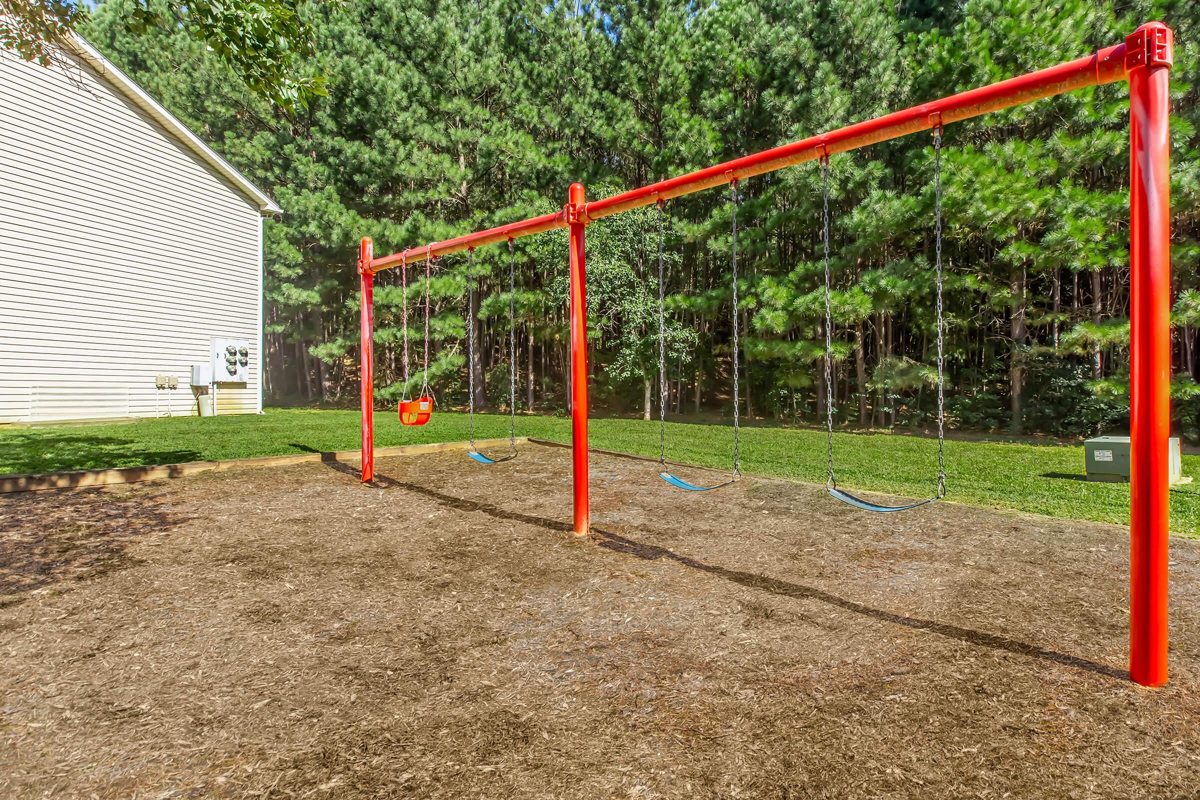 A playground area featuring a red swing set with two blue swings and a red toddler swing. The swings are situated on a dirt surface, surrounded by greenery, with a house partially visible in the background. The scene conveys a tranquil outdoor environment suitable for children's play.