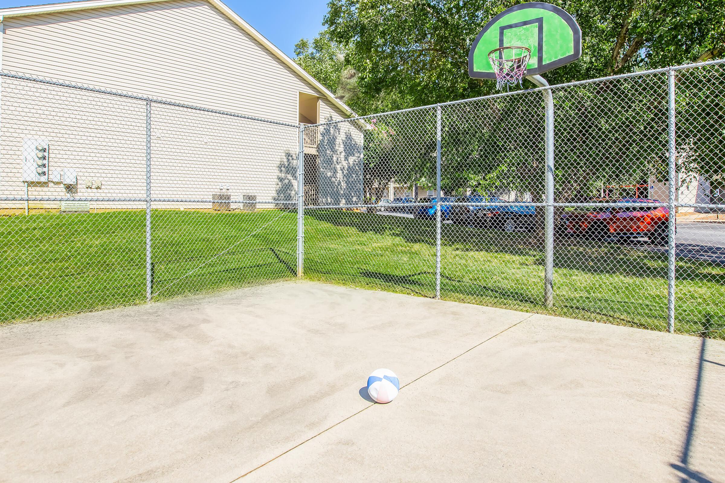A fenced sports area featuring a basketball hoop and a partially deflated soccer ball on the ground. In the background, a grassy area and a building are visible, along with parked cars nearby. The setting is outdoors under bright sunlight.