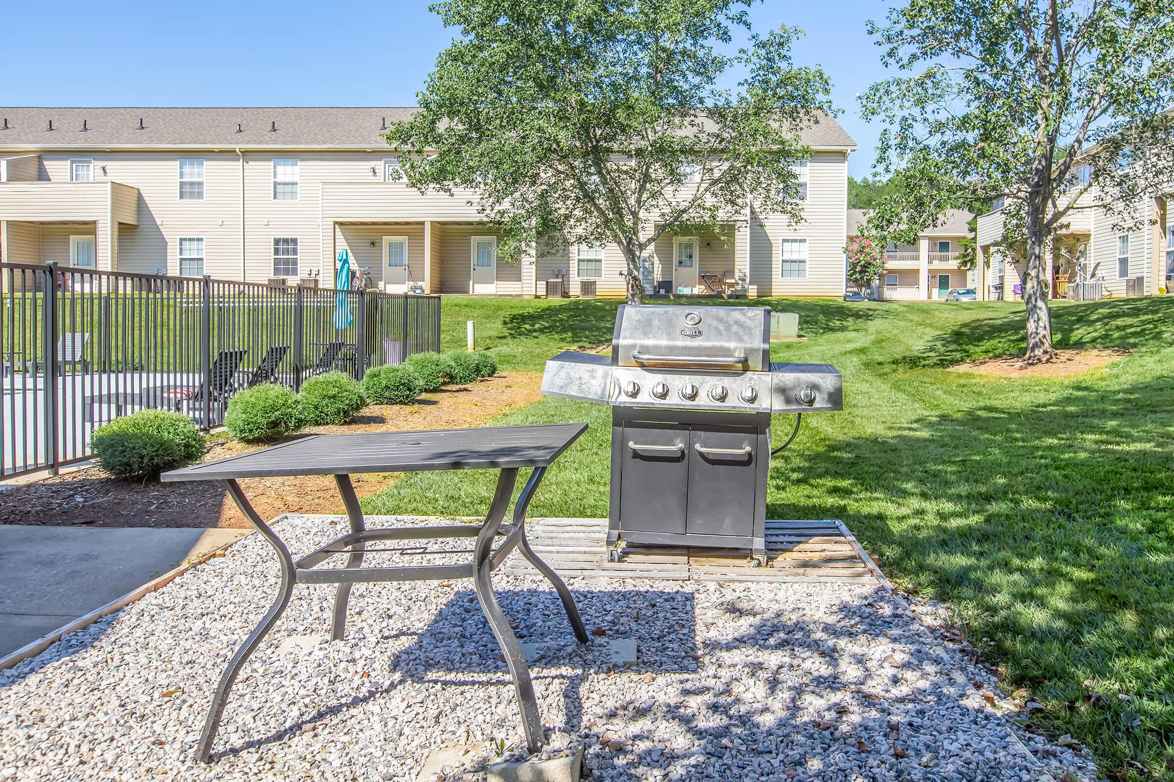 A backyard area featuring a charcoal grill on a stone platform next to a wooden table. In the background, there are several residential buildings and a well-maintained lawn with trees. A fenced pool area is visible nearby, adding to the outdoor recreational space.