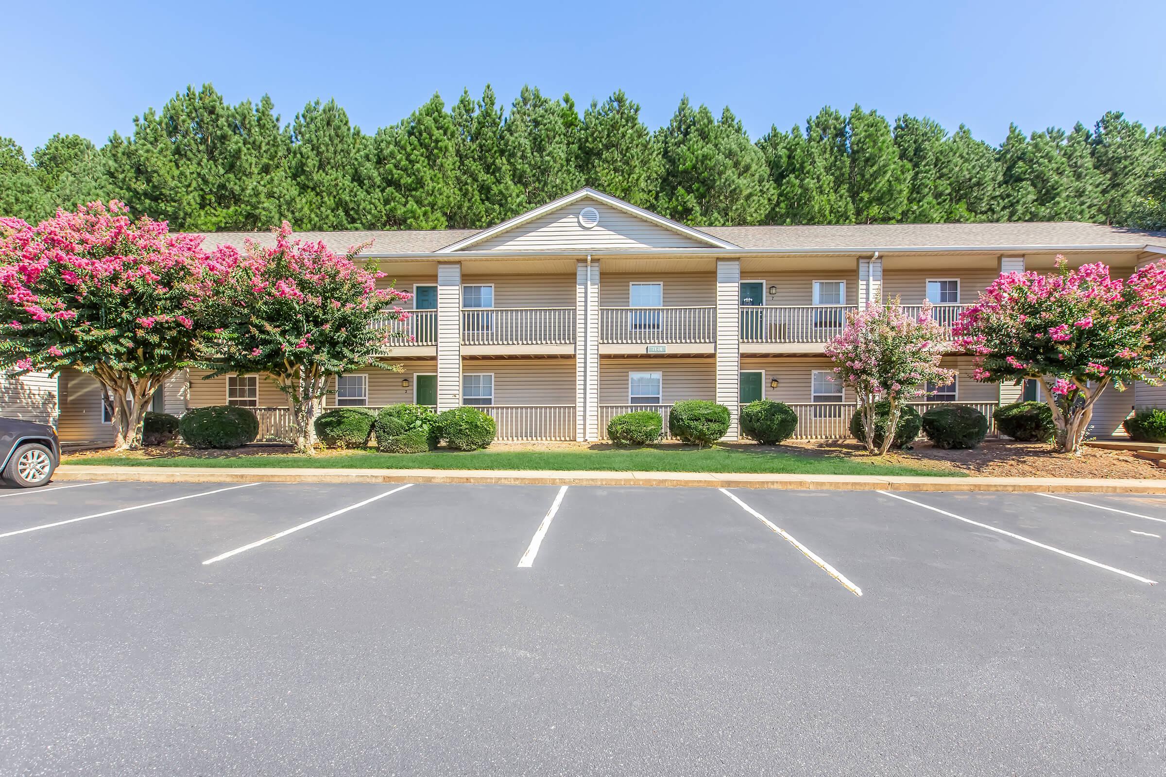 A three-story apartment building surrounded by lush greenery and flowering trees, featuring balconies on each unit. The parking lot in front has several empty spaces, and the clear blue sky enhances the inviting atmosphere of the complex.