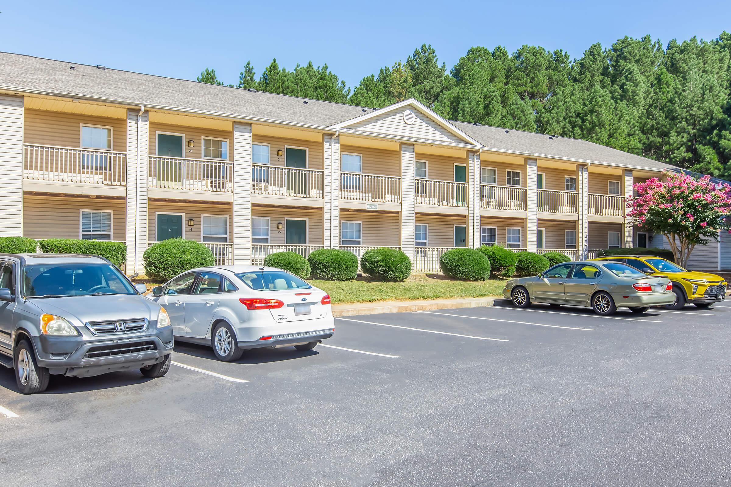 Exterior view of a multi-story apartment building with grassy landscaping and a tree. The building features several balconies and parking spaces occupied by various cars. The setting is surrounded by tall green trees under a clear blue sky.
