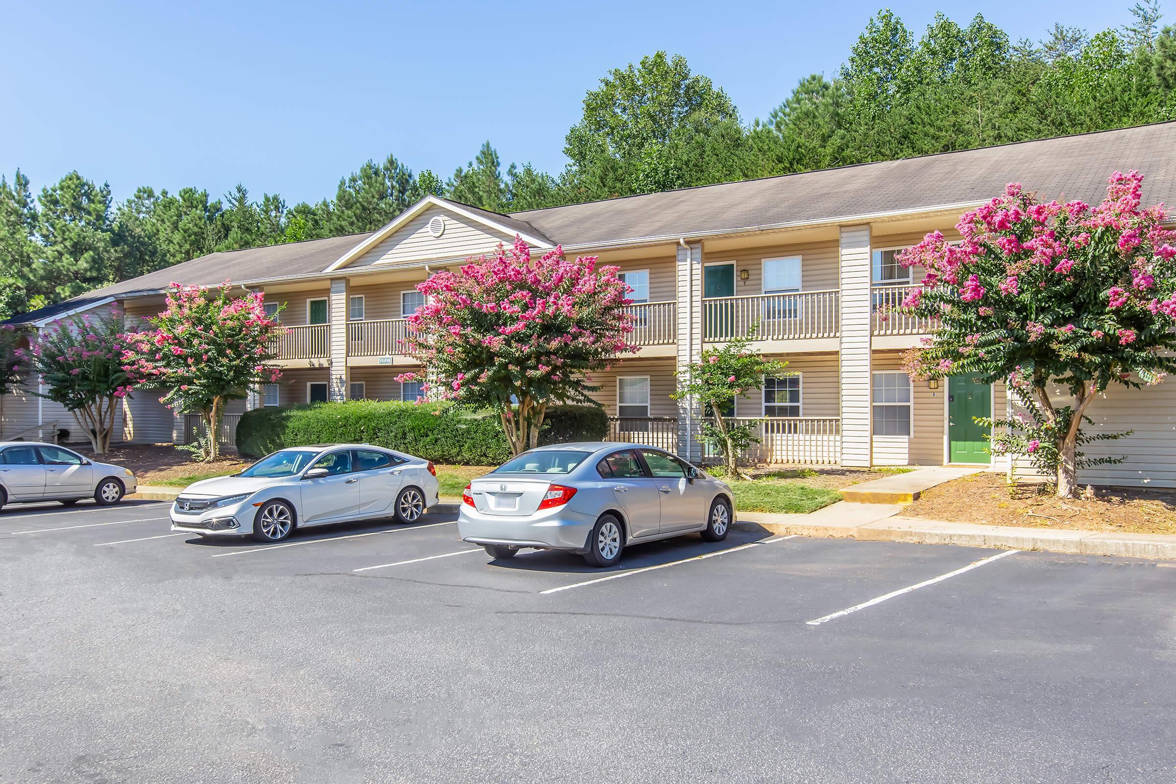 Two-story apartment building with light-colored siding, surrounded by green trees and flowering bushes. Parking lot in front with several parked cars, including a silver sedan and a white car. Clear blue sky in the background.