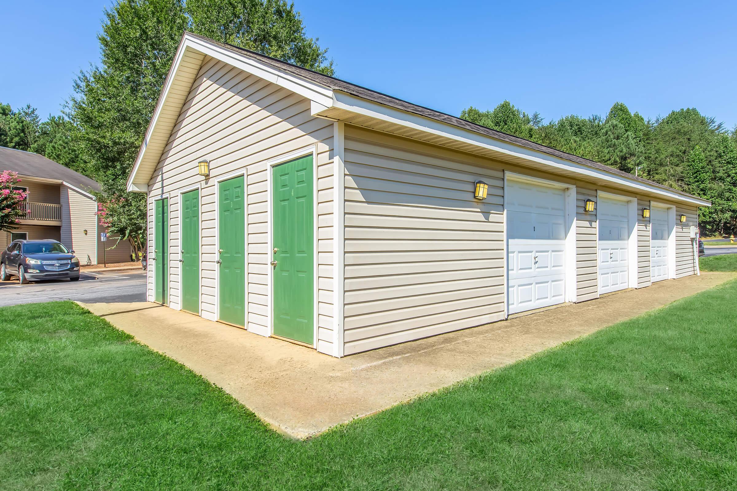 A well-maintained storage unit building featuring multiple green doors on one side, set against a backdrop of trees and clear blue sky. The building has a paved area in front and includes a white garage door on the right. Nearby, there is a parked car and lush green grass surrounding the structure.