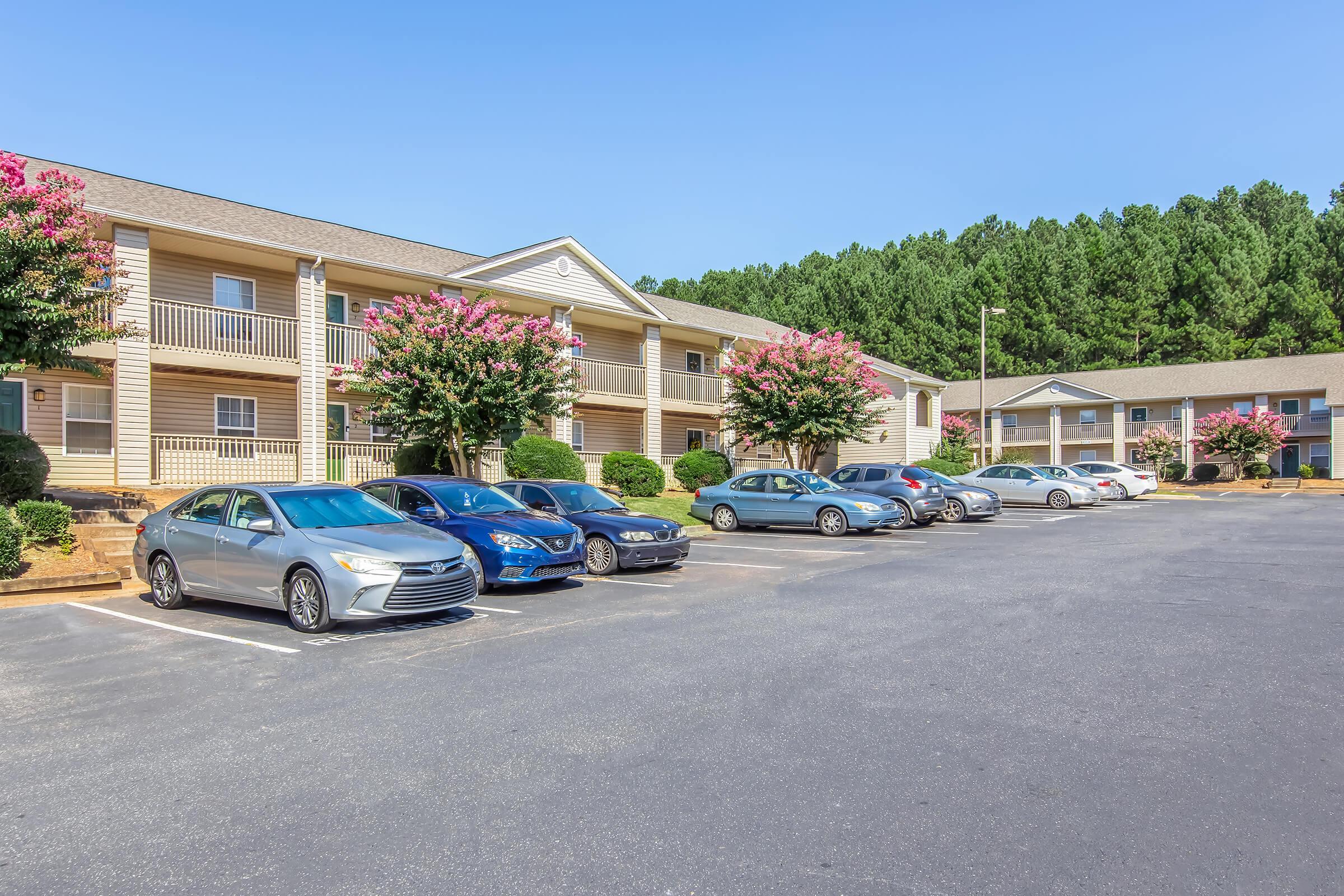 A well-maintained apartment complex with two buildings surrounded by green trees. The foreground features a parking lot with several cars parked in orderly rows. Bright pink flowering bushes add color to the landscape under a clear blue sky.