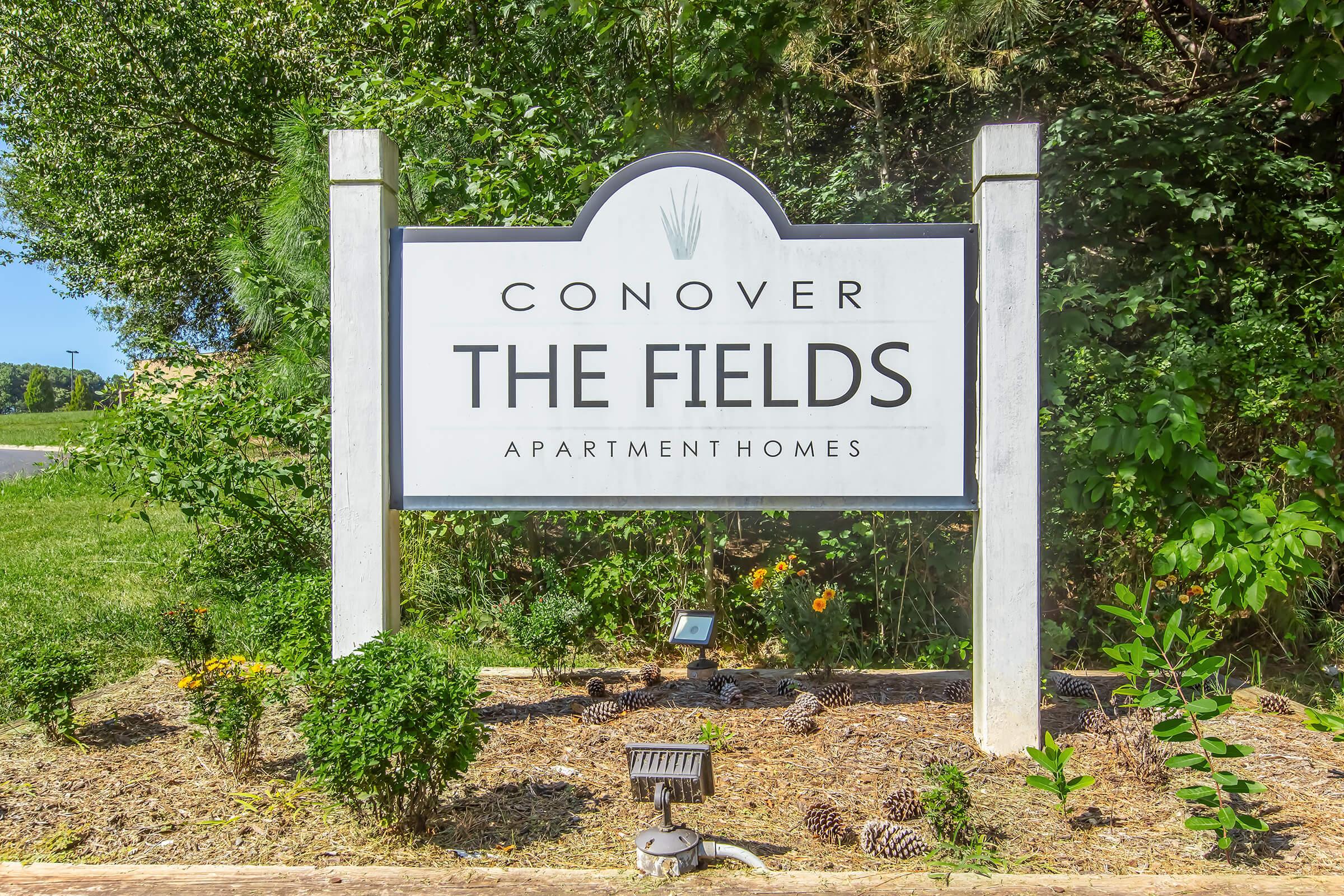 Sign for "The Fields Apartment Homes," with the name prominently displayed along with the word "Conover." The sign is surrounded by greenery, including plants and pinecones, indicating a well-kept outdoor area.
