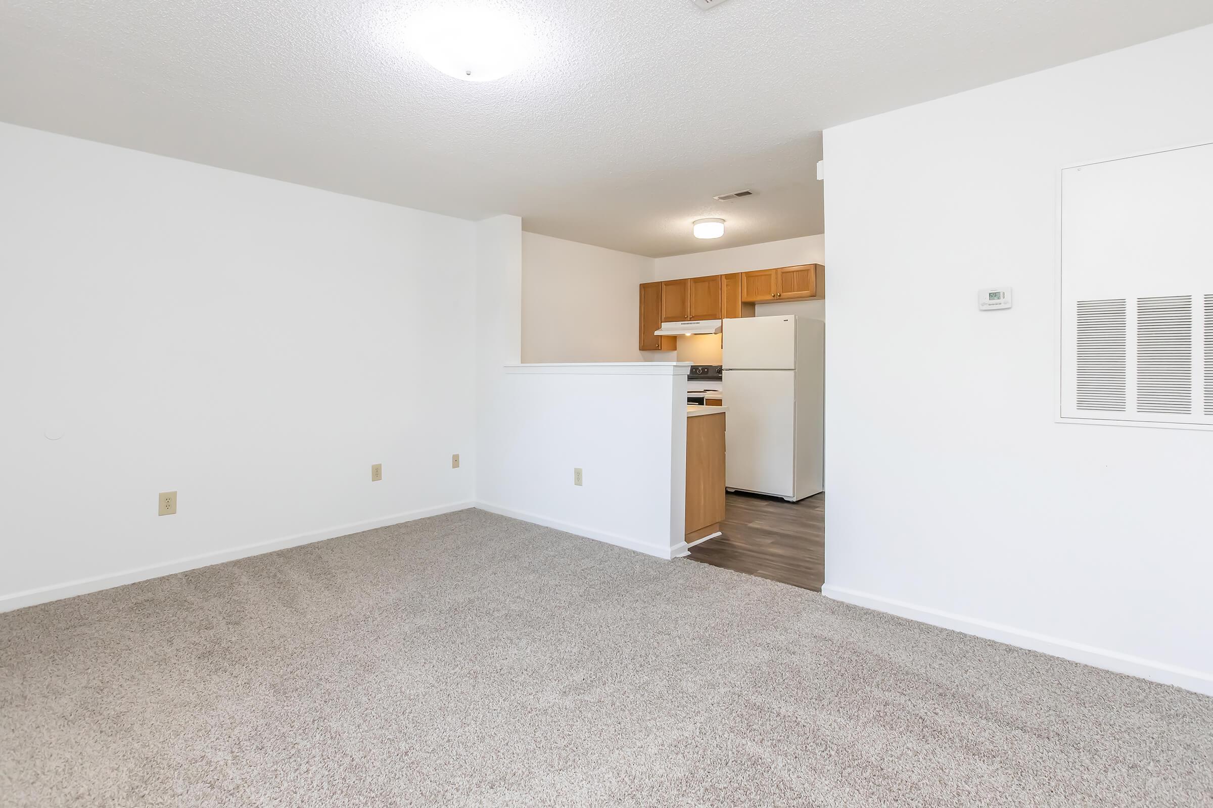 Spacious and clean living area with light beige carpet and white walls. A small kitchen area is visible in the background, featuring wooden cabinets and a white refrigerator. Soft overhead lighting illuminates the room, creating a welcoming atmosphere.