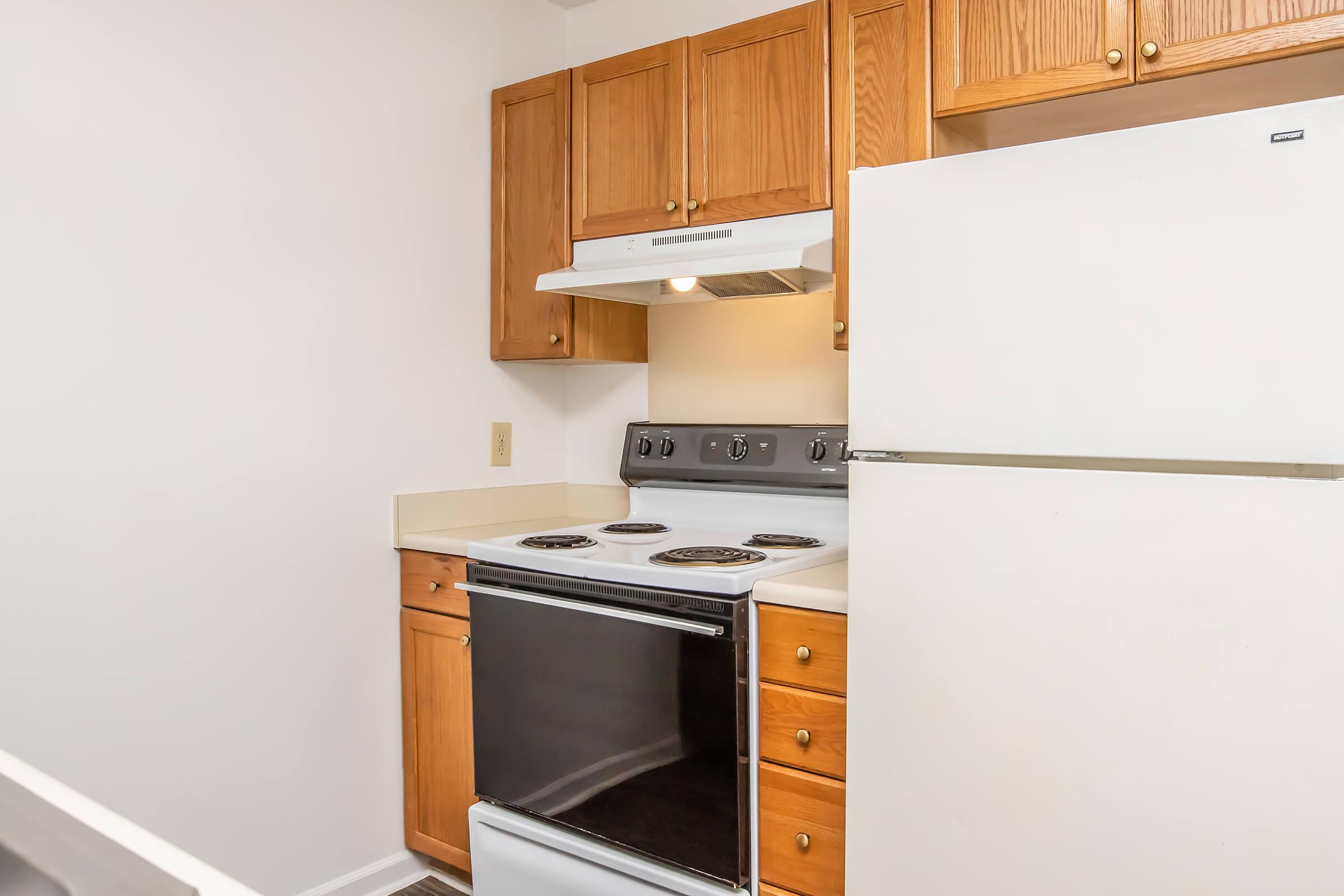 A small kitchen featuring light wooden cabinets, a white refrigerator, and a black stove with four burners. The countertop is beige and there is an exhaust hood above the stove. The walls are painted a light color, creating a bright, open feel.