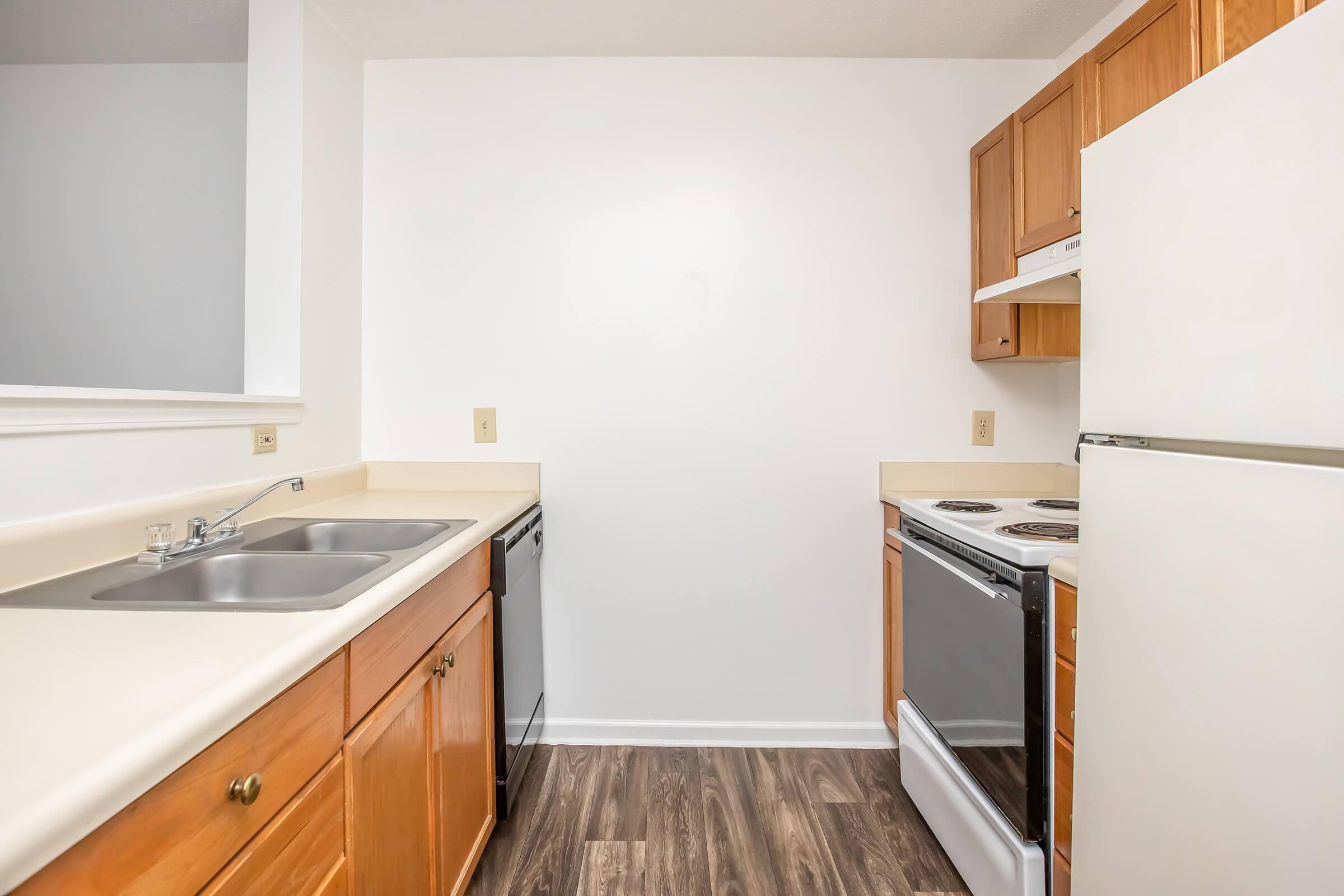 Small kitchen featuring wooden cabinets, a double sink, a dishwasher, and a white refrigerator. The kitchen includes a stove and oven combo on the right, with a light-colored countertop and laminate flooring. The walls are painted white, giving the space a bright and clean appearance.