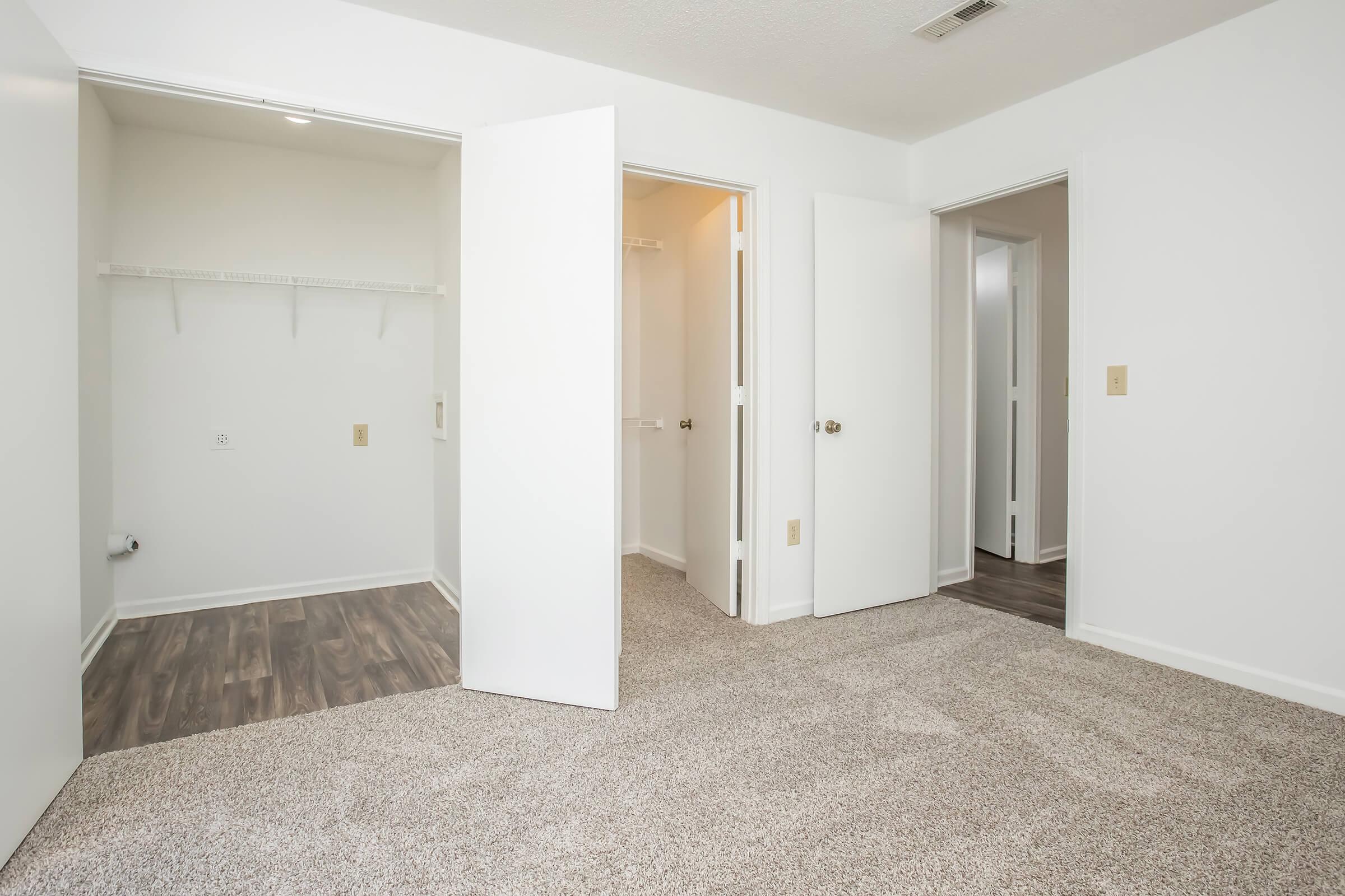 A bright, empty room with light-colored walls and neutral carpeting. Two open doors lead to closets, while a third door is visible in the background. The space is well-lit and tidy, suggesting a fresh and inviting atmosphere, suitable for various uses such as a bedroom or office.