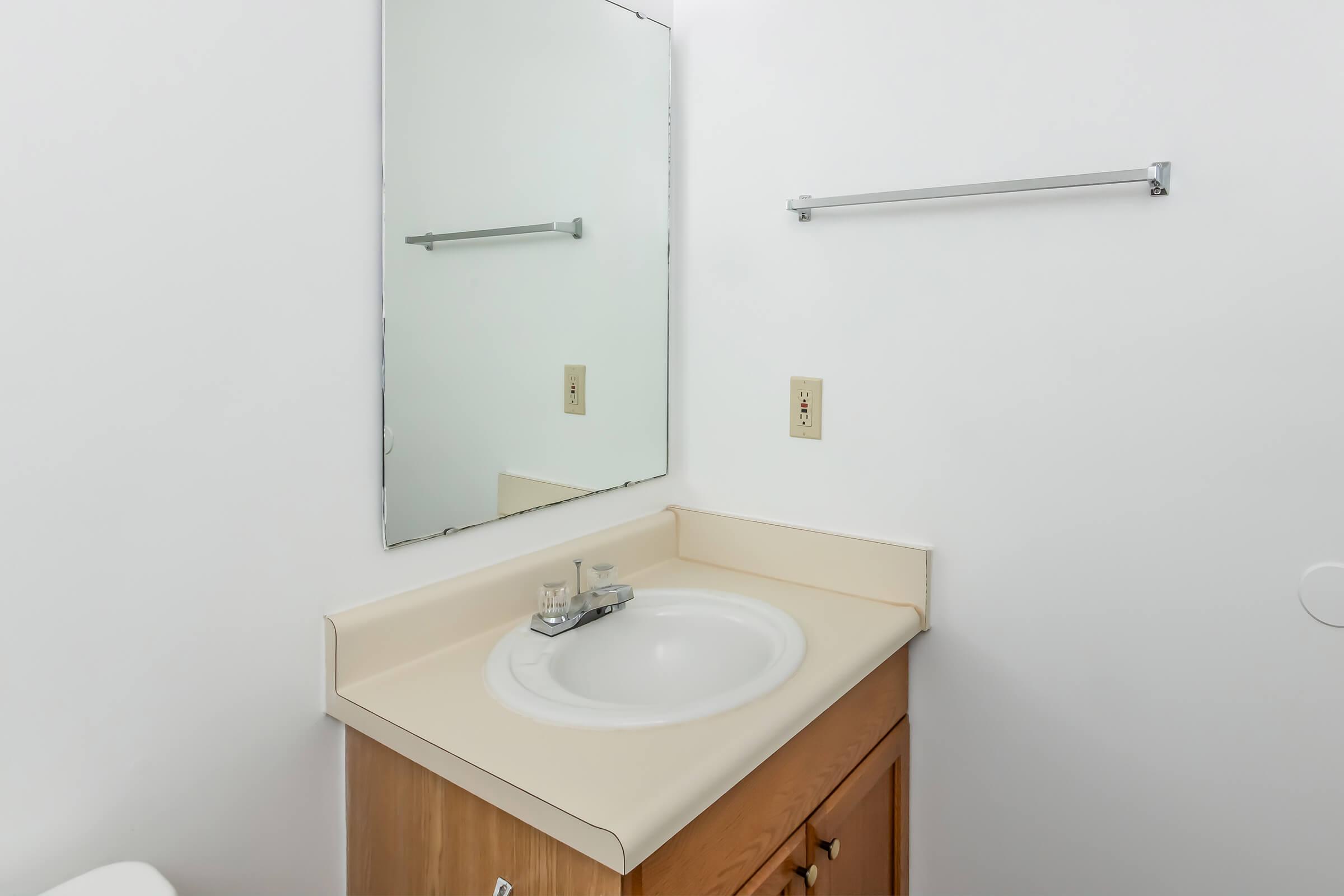 A simple bathroom sink area with a beige countertop and a mirror above. The sink features a faucet and sits on a wooden cabinet. A towel bar is mounted on the wall next to the mirror, and the walls are painted white, giving a clean and minimalist look.