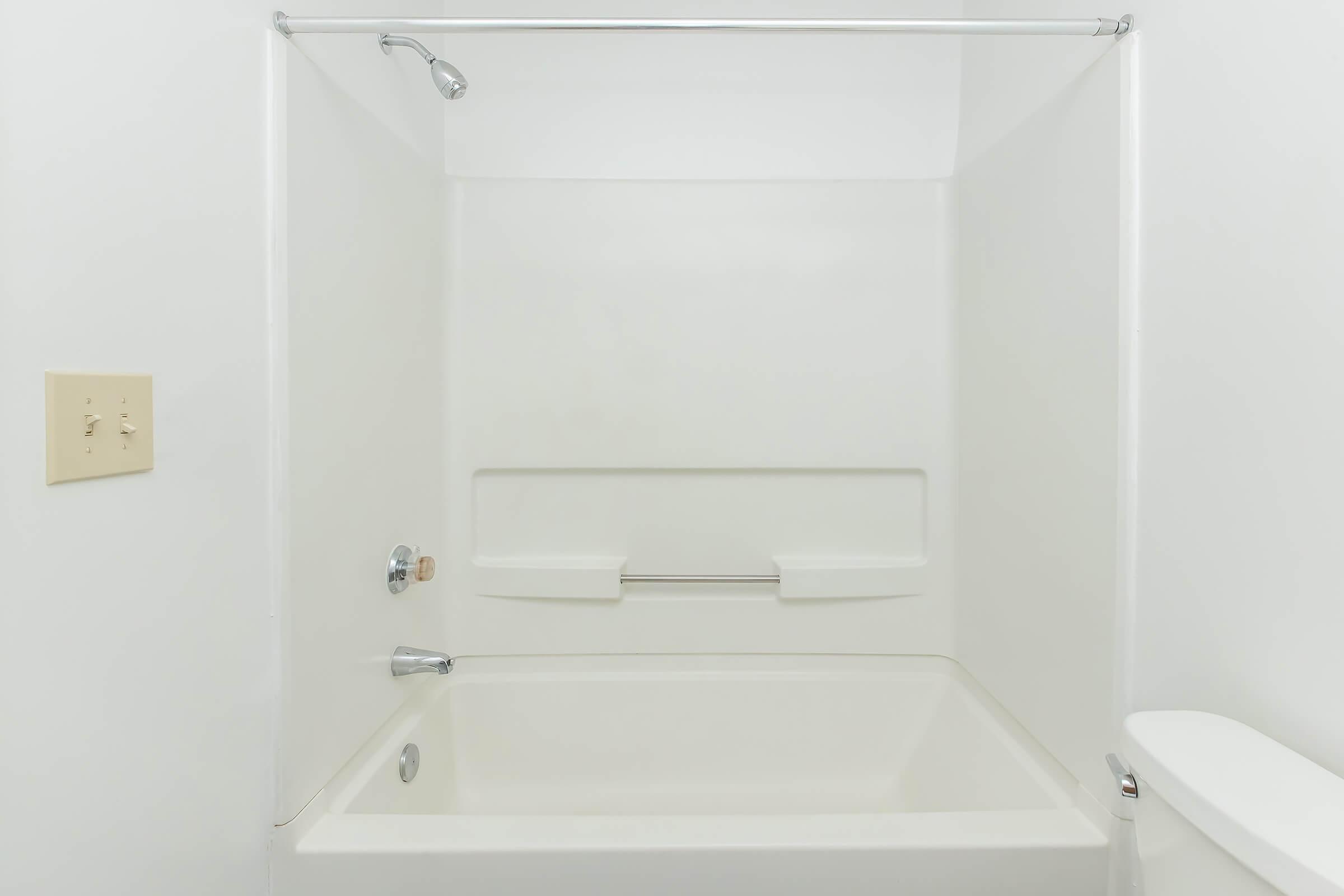A clean, empty bathroom with a white bathtub, a showerhead mounted above, and a grab bar on the wall. The walls are plain white and there is a toilet visible to the right. The overall ambiance is simple and minimalistic.
