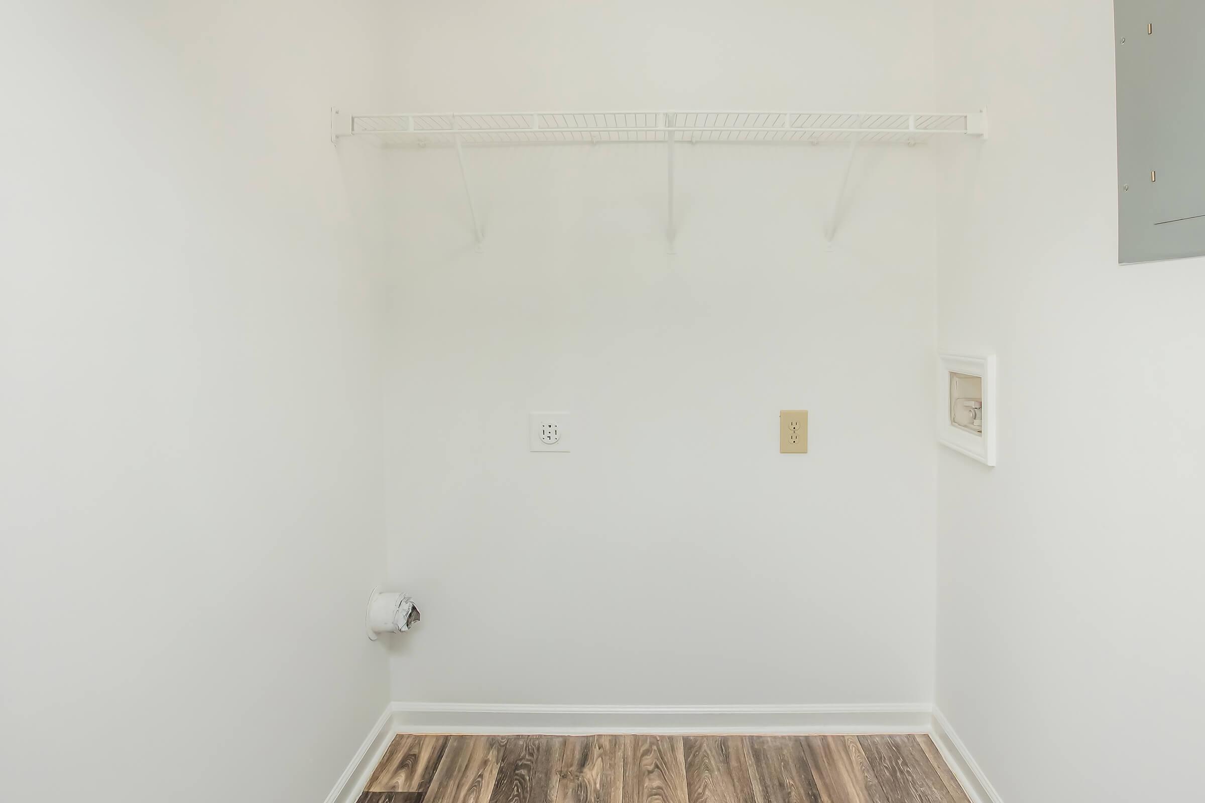 Empty room with white walls and a wooden floor. A wire shelf is mounted on the wall, and there are two electrical outlets visible. A small section of the wall on the right has a panel, and there is a visible duct or vent at the bottom left. The room appears to be a laundry or storage area.