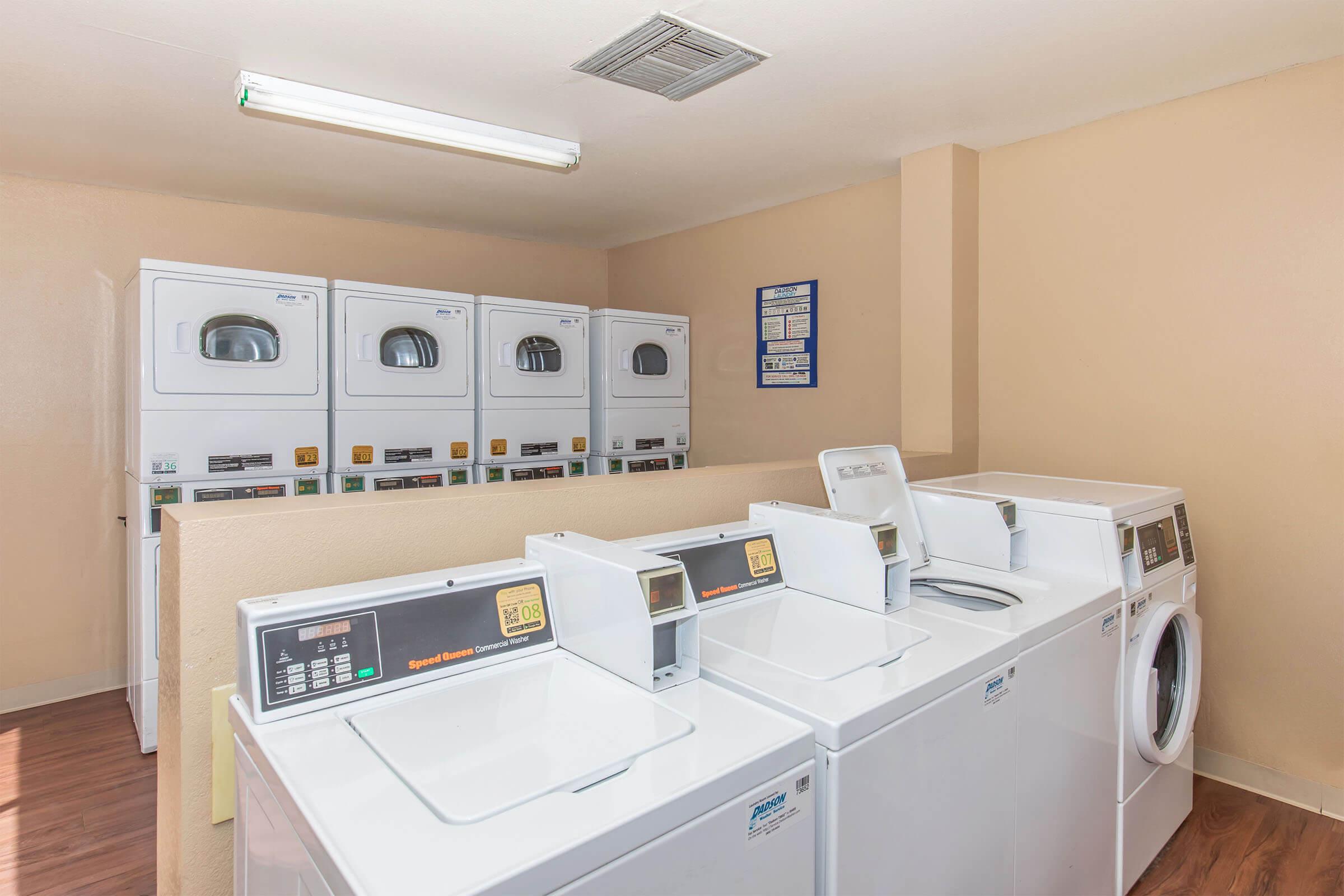 A clean laundry room featuring several rows of coin-operated washing machines and dryers. The walls are painted in a light shade, and there is a notice board with laundry instructions. Soft lighting illuminates the space, creating a functional and inviting atmosphere for guests.