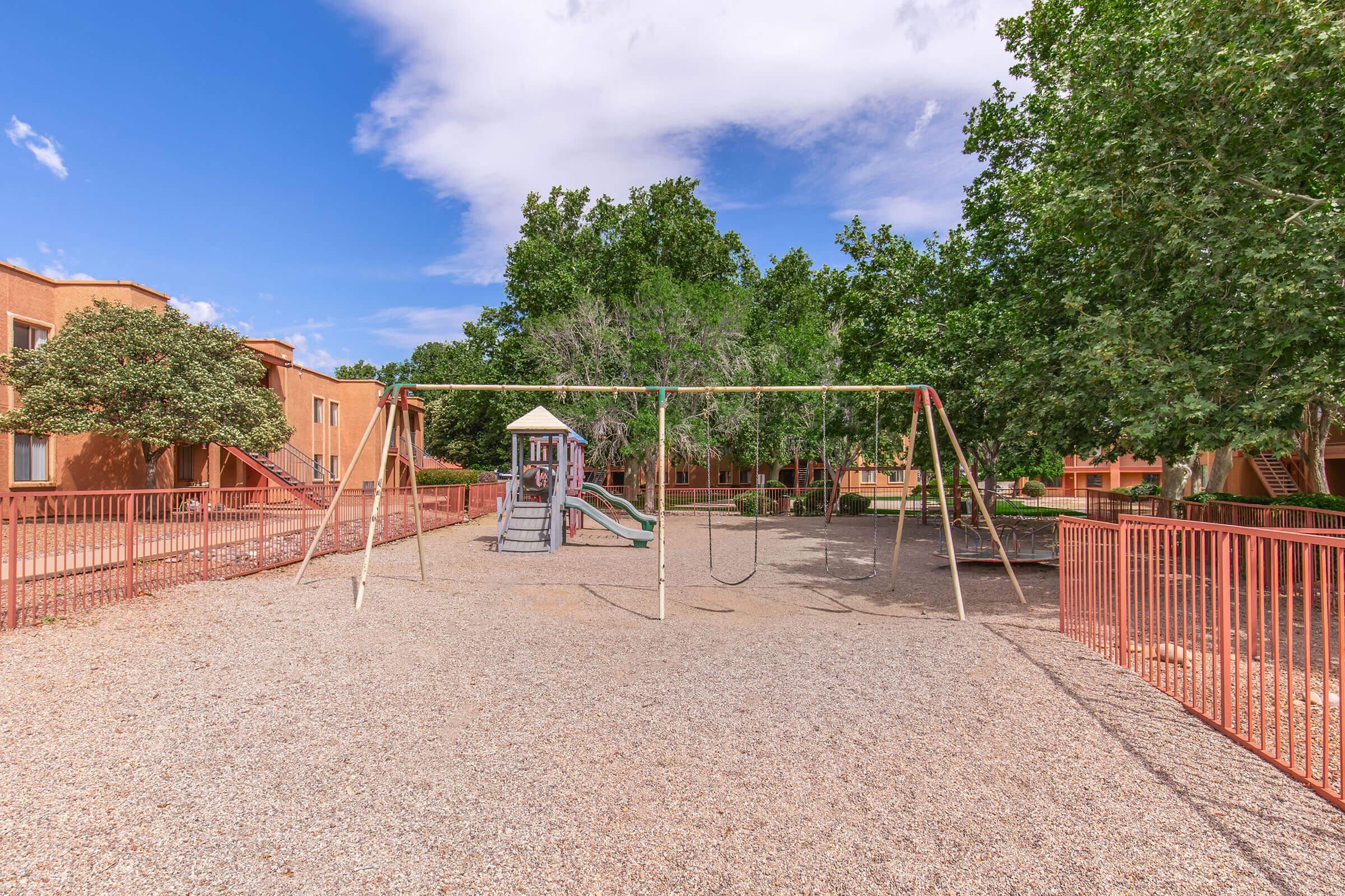 A playground area featuring swings and a slide, surrounded by a gravel surface and several trees. The backdrop includes apartment buildings with an orange hue, under a blue sky with scattered clouds.
