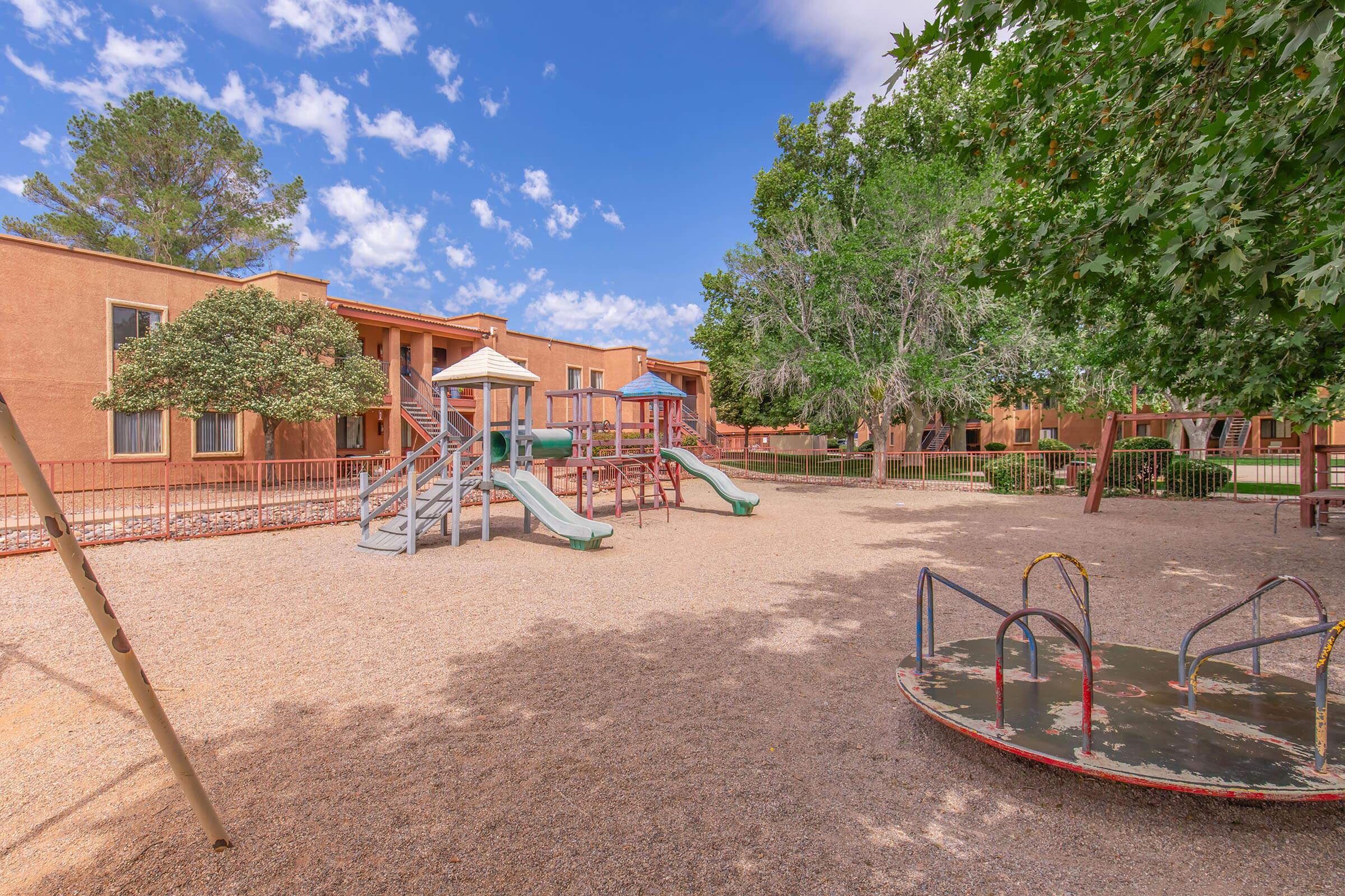 Playground scene featuring colorful slides and a merry-go-round in a sandy area. Surrounding trees provide shade, and there are residential buildings in the background under a bright blue sky with scattered clouds. The setting is inviting for children.