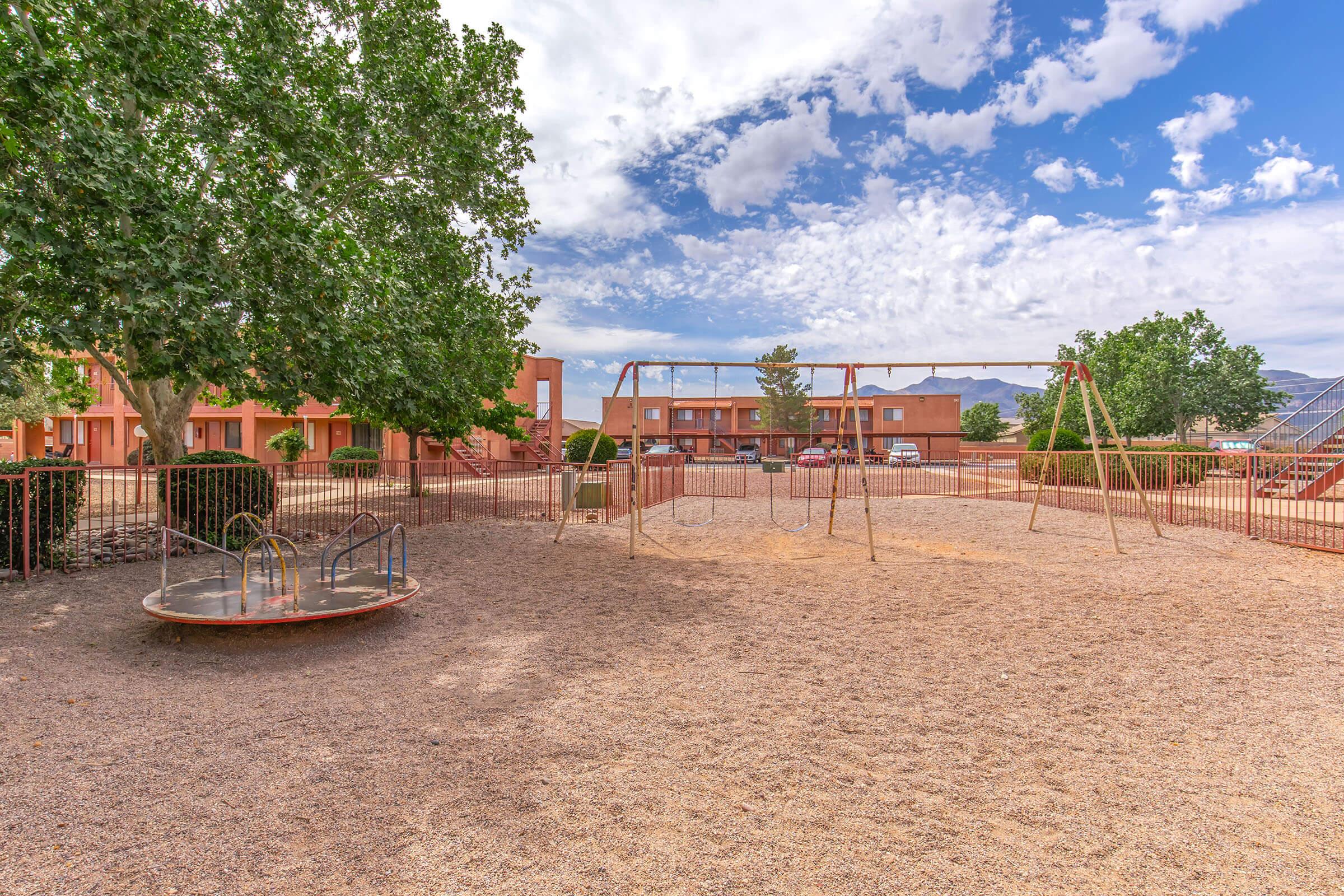 A playground scene featuring swings and a roundabout, set in a sandy area surrounded by trees. In the background, there are buildings with a cloudy blue sky above. The playground equipment is unoccupied, suggesting a peaceful environment ideal for children.