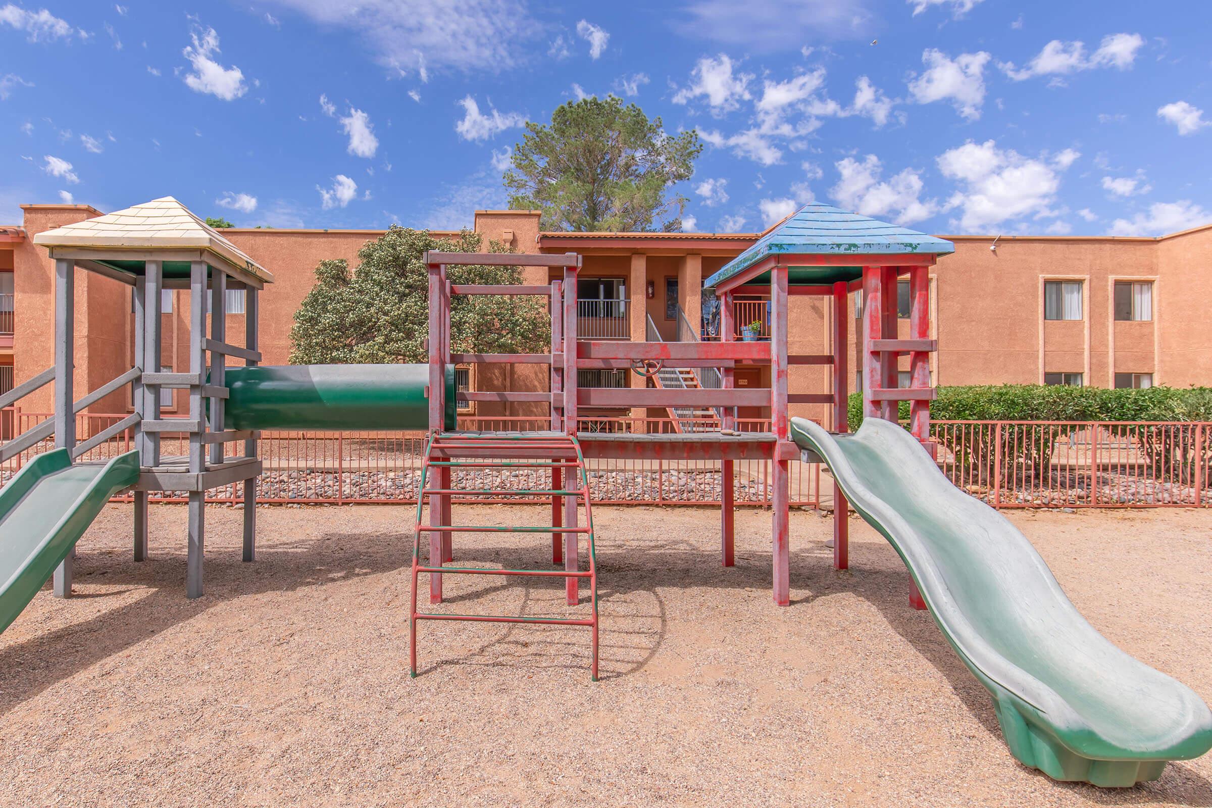 A colorful playground featuring two play structures with slides, surrounded by a sandy area and bordered by a fence. In the background, there are residential buildings and a clear blue sky with some clouds.