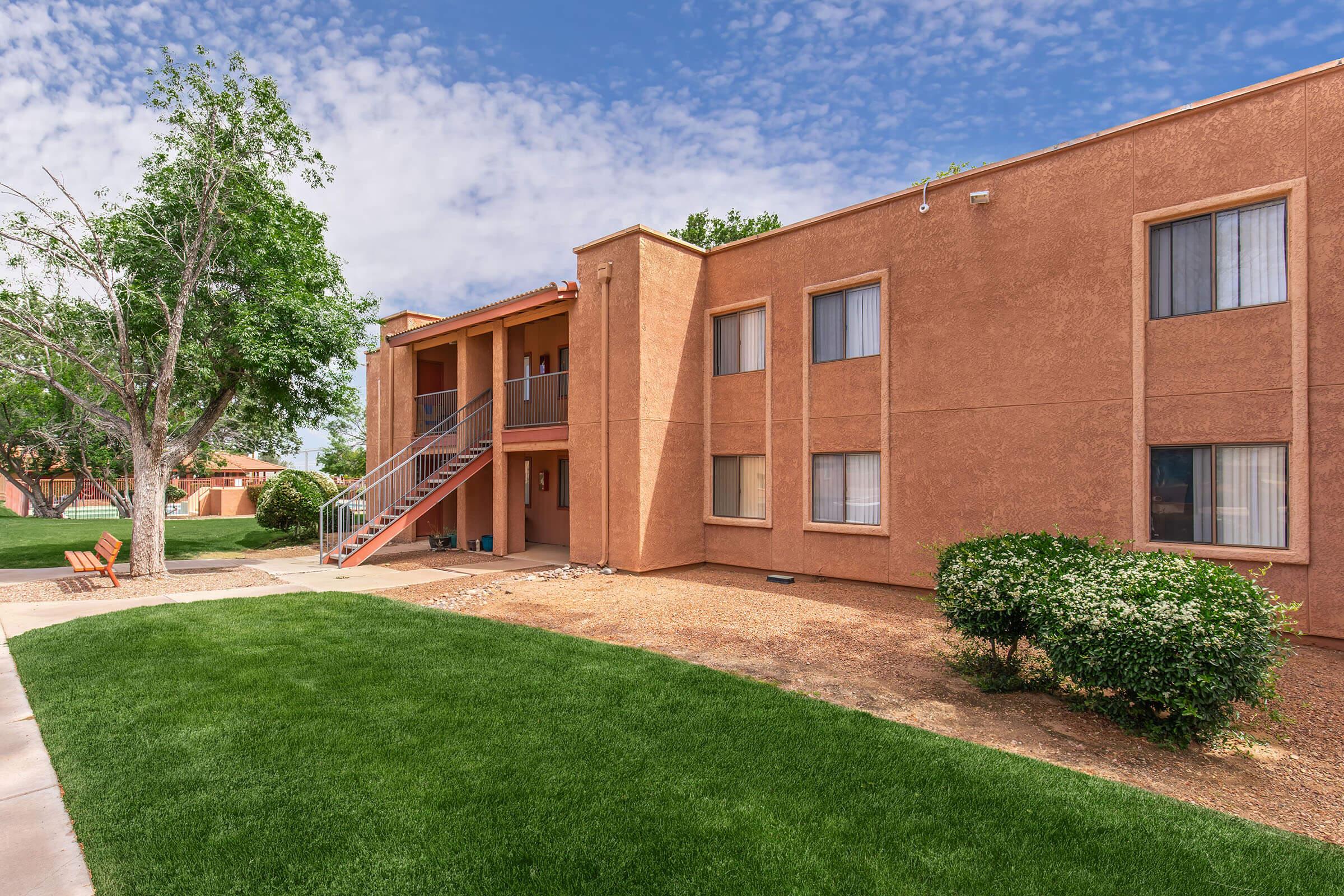 A two-story apartment building with a brown exterior, featuring a staircase leading to the upper floor. The well-maintained lawn surrounds the building, and there are a few trees and shrubs. The sky is partly cloudy, adding a touch of blue to the scene.