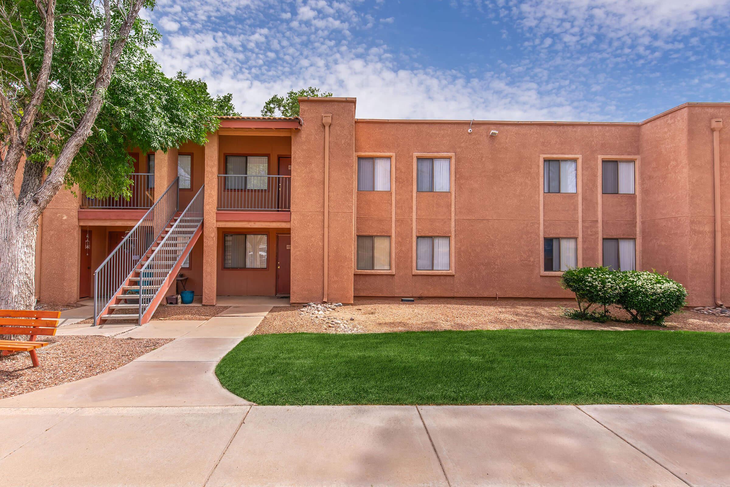 Exterior view of a two-story peach-colored apartment building. The entrance features a set of stairs leading to the second floor, with green trees nearby. A small patch of grass and rocks adds to the landscape, while a bench sits along the walkway. The sky is partly cloudy, creating a bright atmosphere.