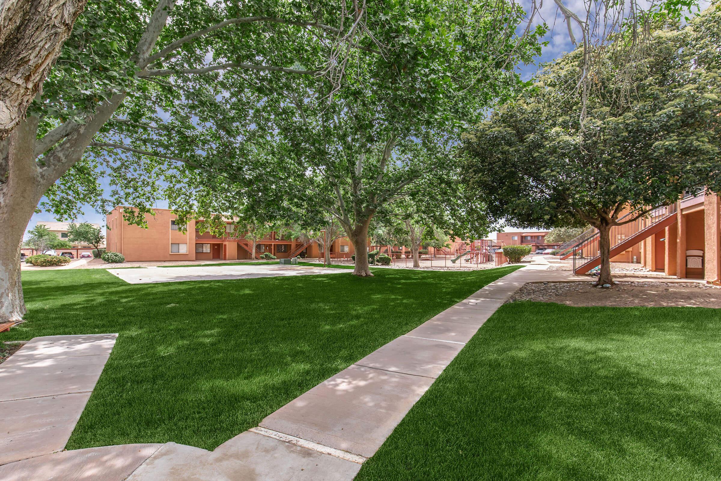 Green lawn with well-maintained grass, lined with concrete pathways. Large trees provide shade, surrounding a multi-story apartment complex with reddish buildings in the background. The scene is bright and sunny, conveying a peaceful residential atmosphere.