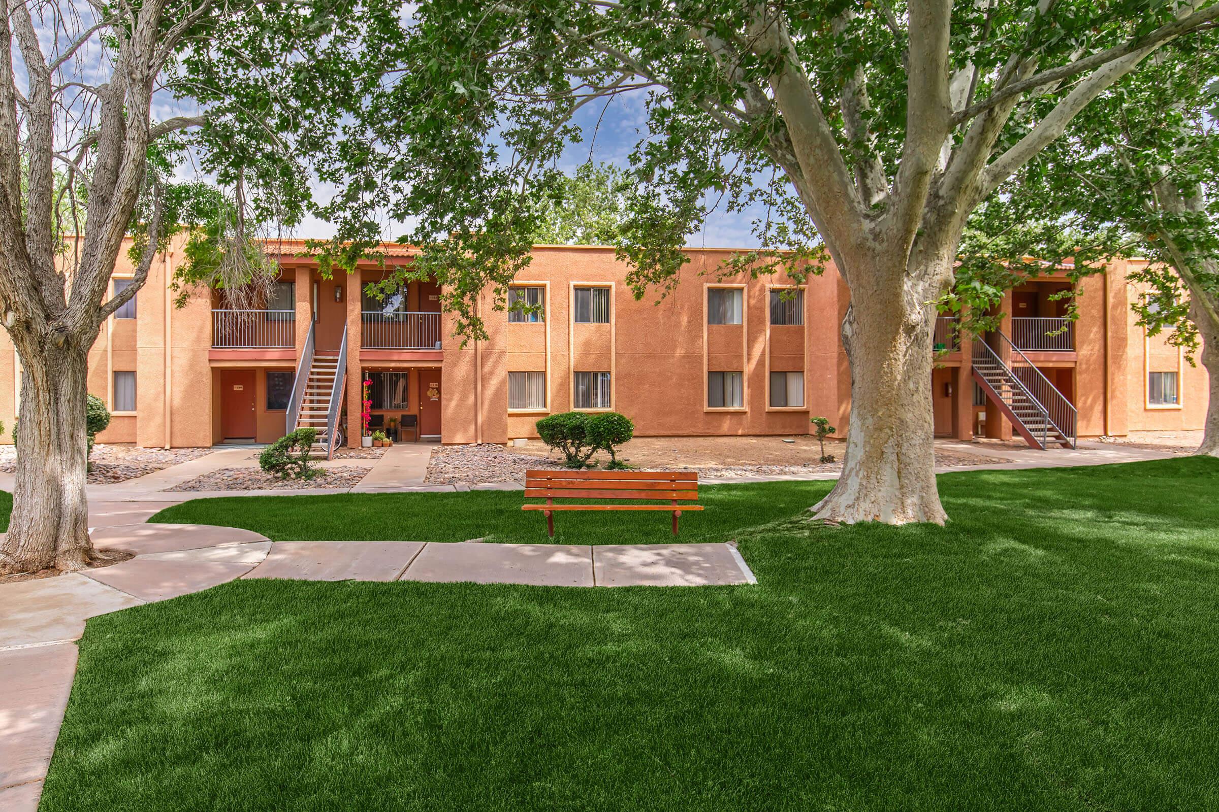 A well-maintained apartment complex with a warm orange exterior, surrounded by green grass and neatly trimmed trees. The image features a central pathway leading to a wooden bench and two staircases leading to upper-level units. The sky is clear, adding to the inviting atmosphere of the outdoor space.