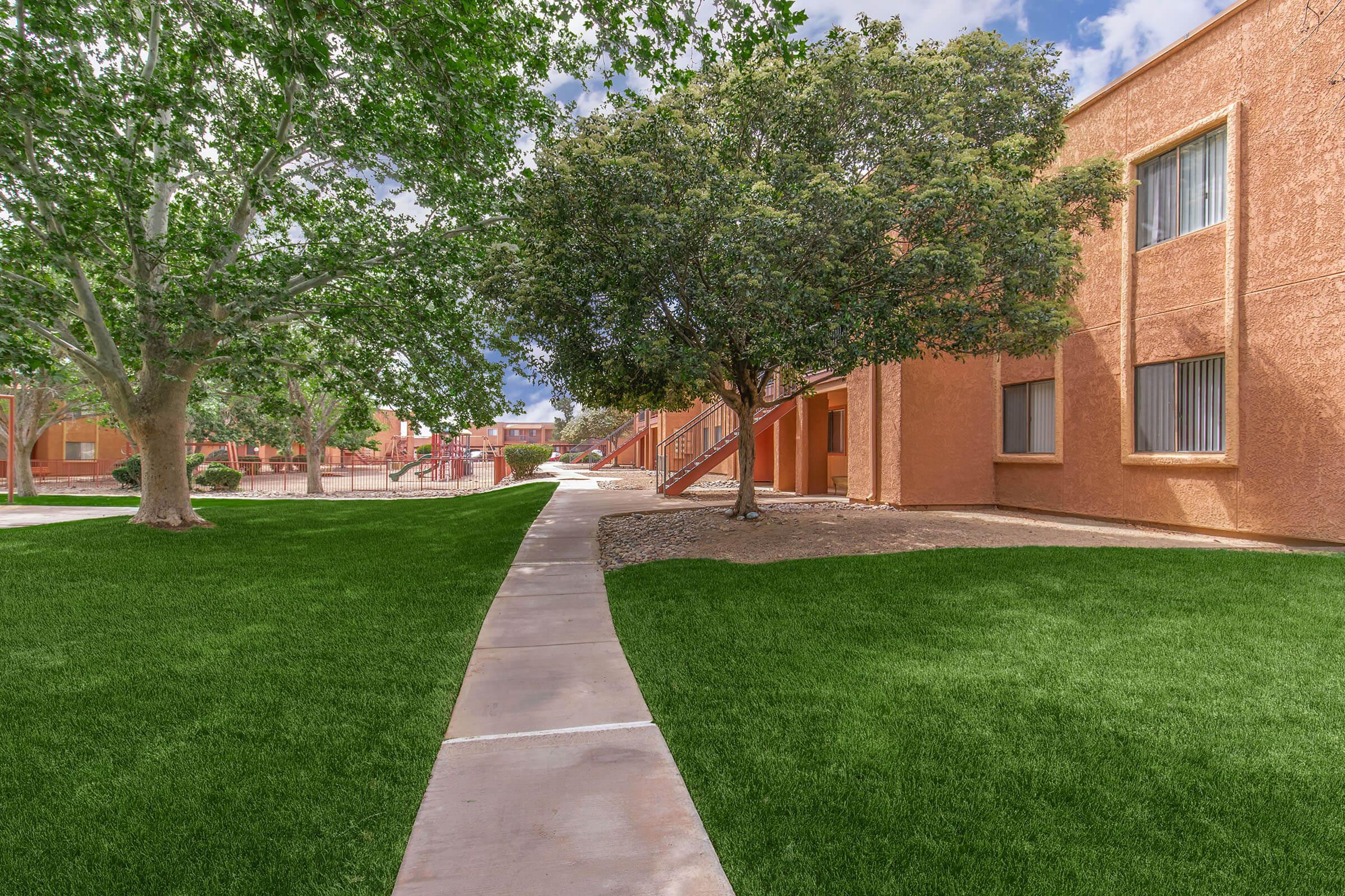 Lush green lawn with a concrete walkway leading through trees towards an apartment complex. The buildings are a warm orange hue, and the sky is partly cloudy, creating a bright and inviting outdoor scene.