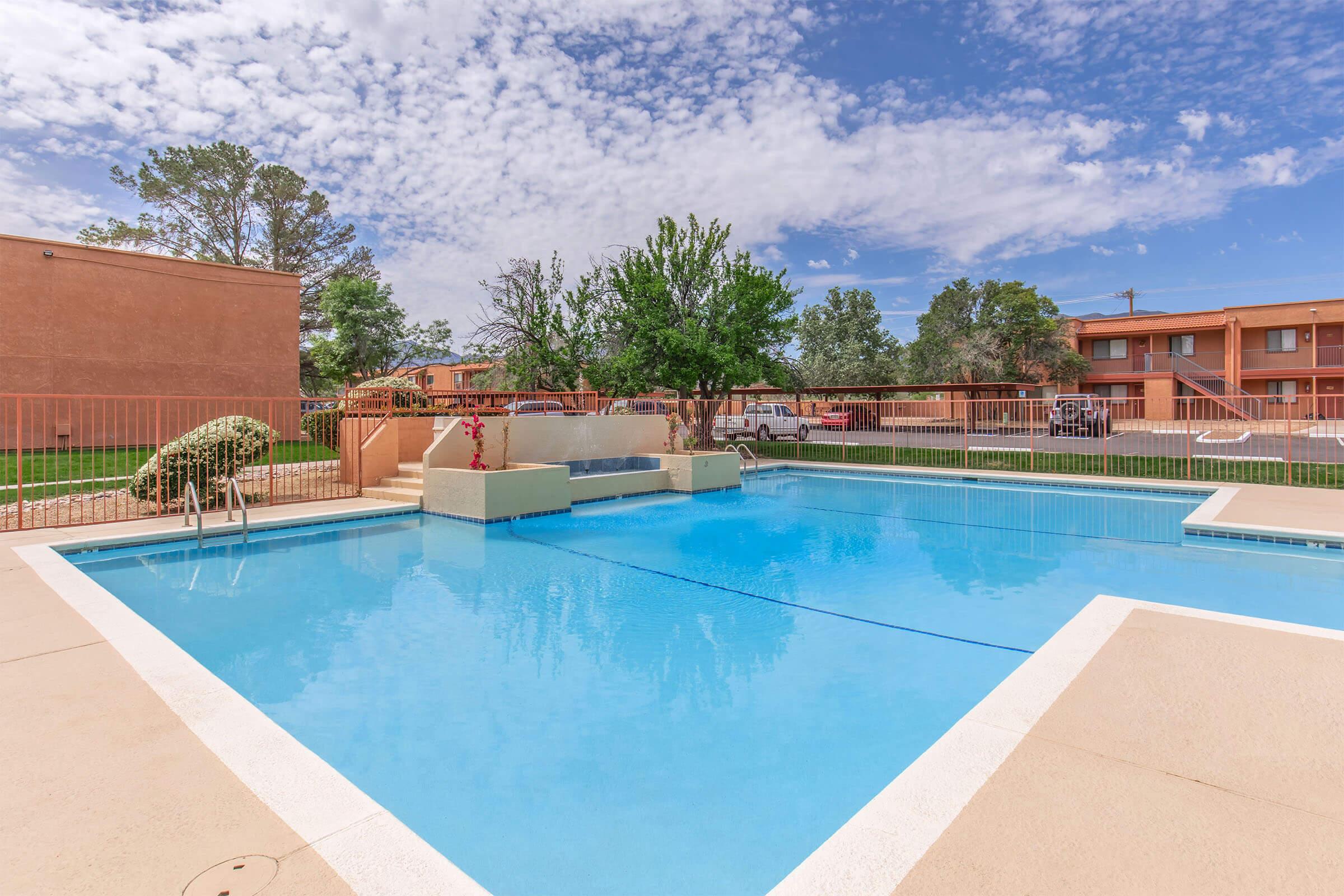 A clear, blue swimming pool surrounded by a tan patio. There are lounge chairs positioned nearby, with a fence enclosing the area. In the background, there are two multi-story buildings with balconies. The sky is partly cloudy with a few trees visible around the pool area.