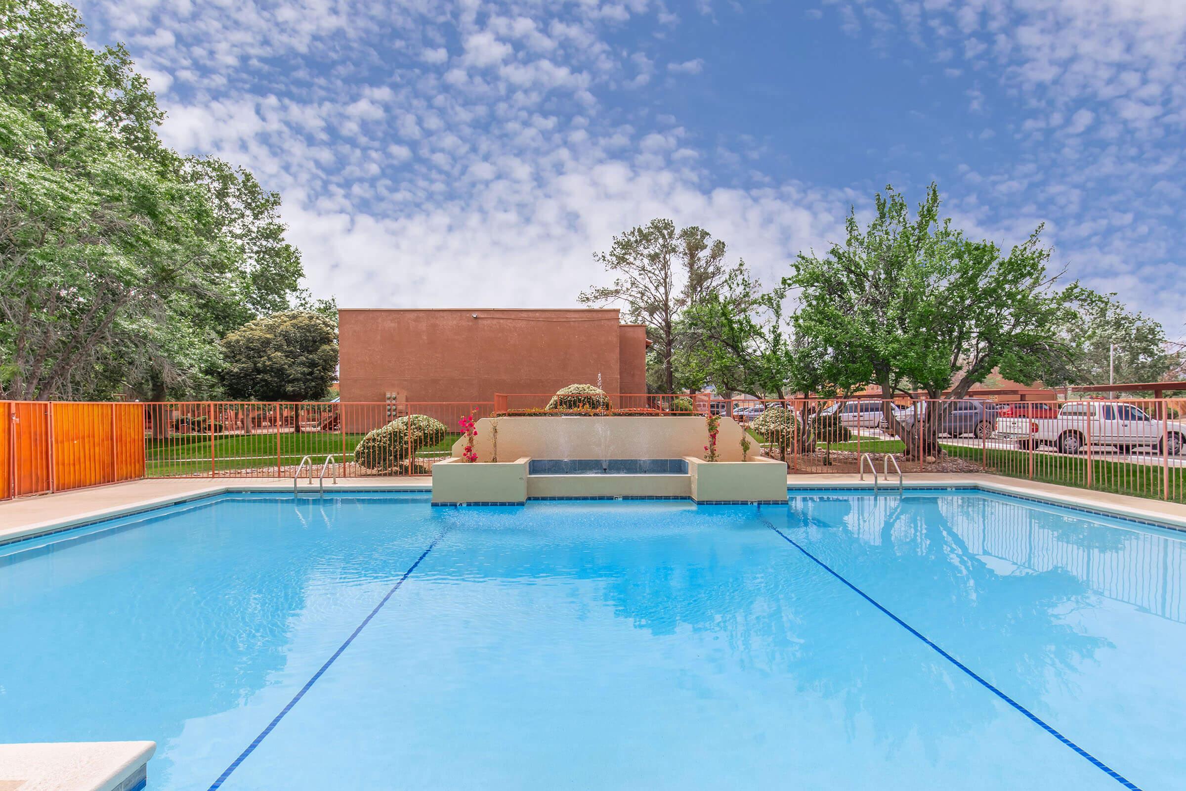 A clear blue swimming pool with a central fountain feature, surrounded by a spacious deck and lounge chairs. Green trees and shrubbery line the background, with a wall and building visible in the distance under a cloudy blue sky.