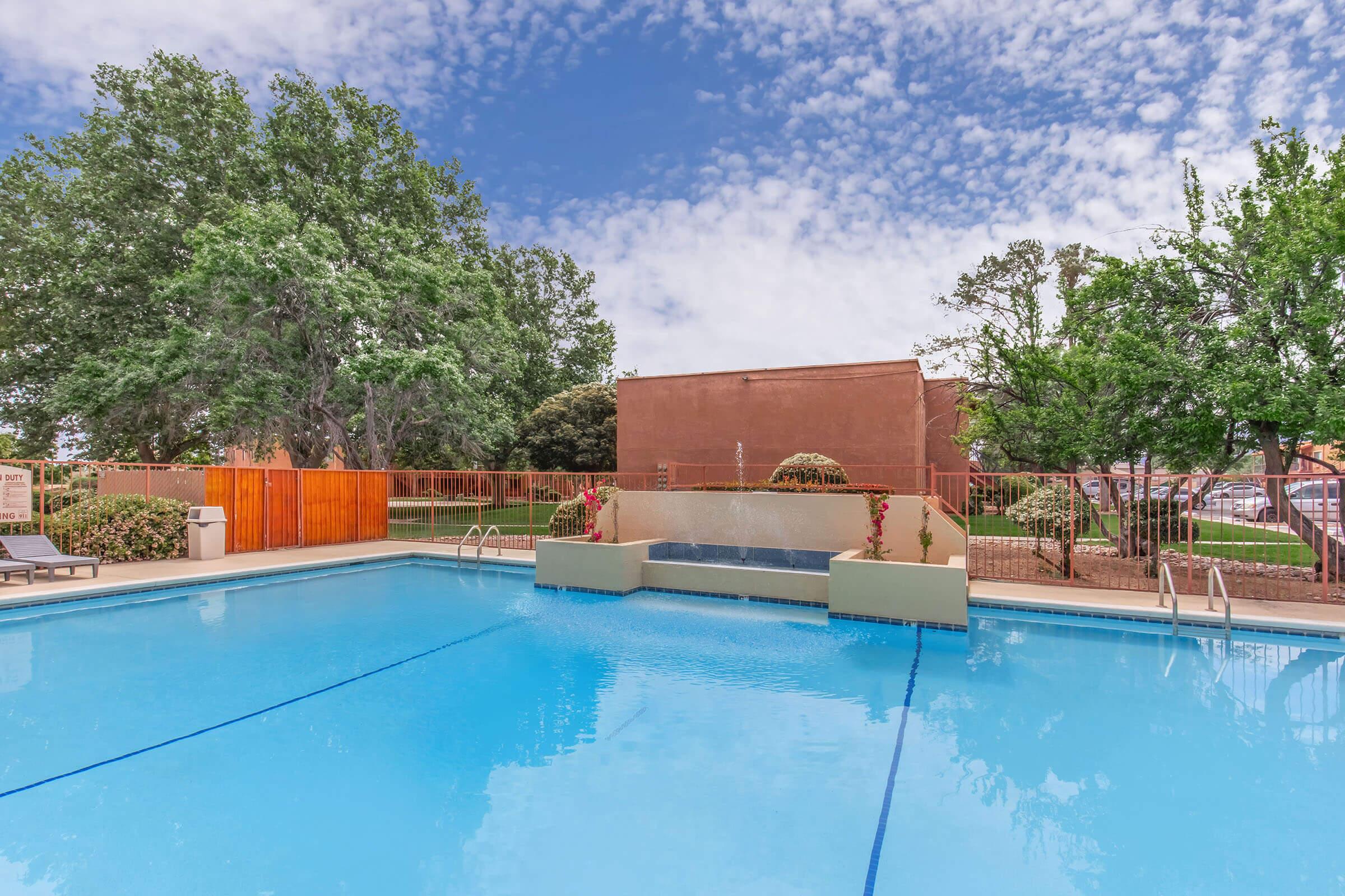 A clear swimming pool surrounded by lush greenery and a decorative wall. The pool features a waterfall, and the sky is filled with scattered clouds. There are lounge chairs at the pool's edge, providing a relaxing atmosphere. The background includes trees and a wooden fence.