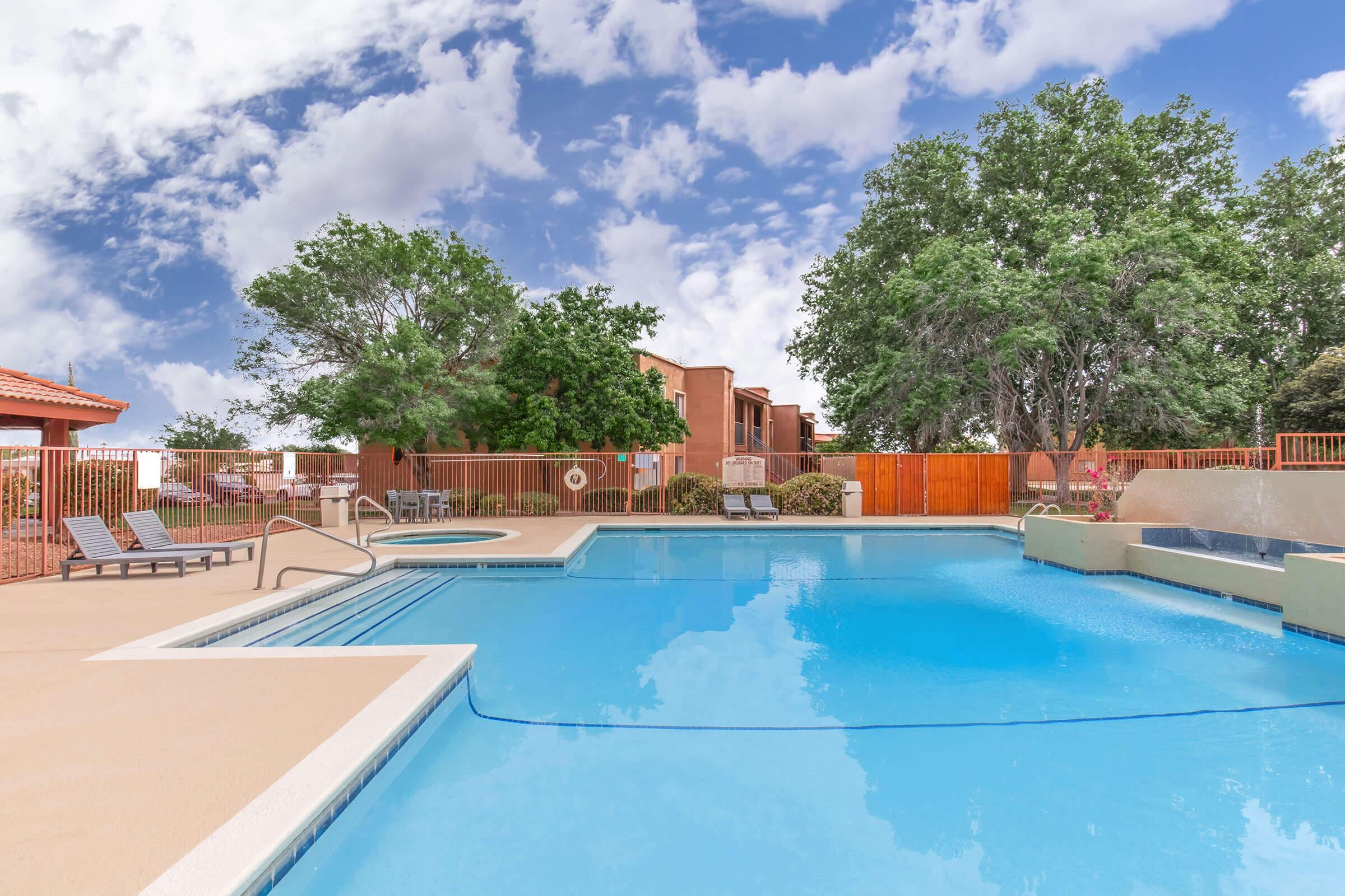A clear blue swimming pool with lounge chairs on the side, surrounded by greenery and a wooden fence. In the background, there are residential buildings and a partly cloudy sky, creating a relaxing outdoor atmosphere.