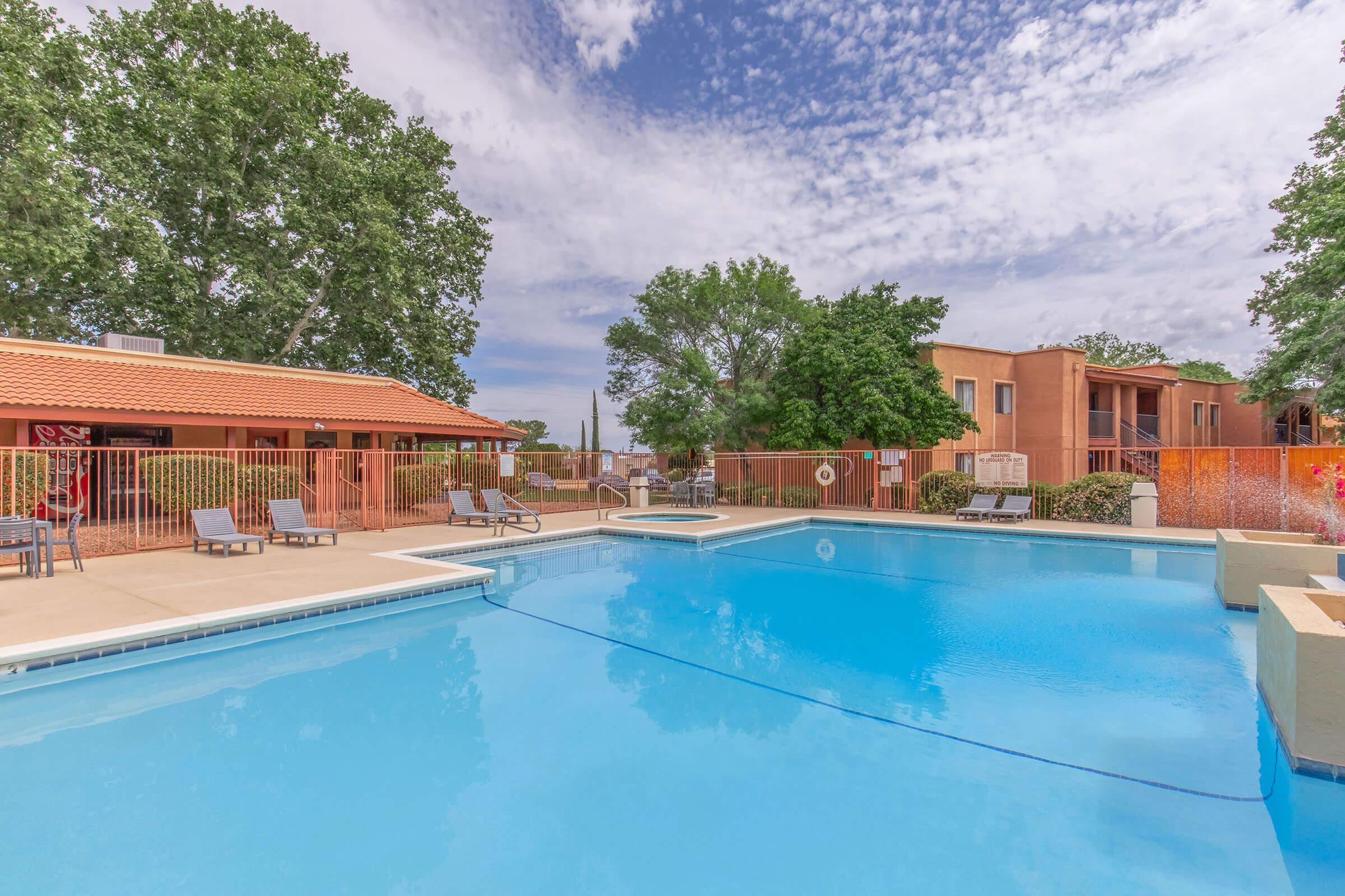 A bright, sunny outdoor pool area featuring a large swimming pool with clear blue water, surrounded by lounge chairs. In the background, there are buildings with reddish-brown stucco exteriors and trees, under a partly cloudy sky. The scene conveys a relaxing atmosphere suitable for leisure.