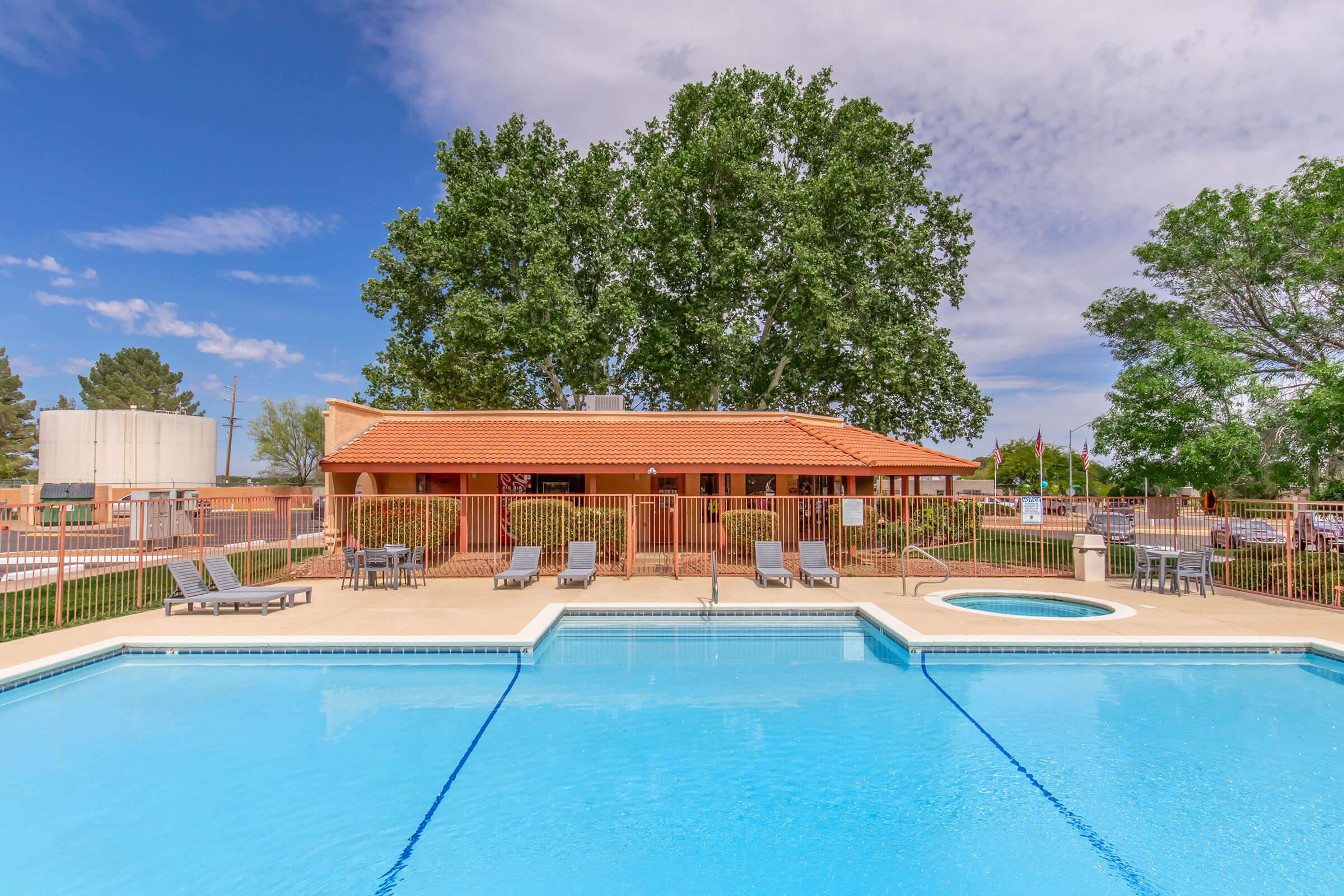 A clear blue swimming pool with a hot tub, surrounded by lounge chairs. In the background, a single-story building with a red tile roof is visible, along with green trees and a blue sky dotted with clouds. The area appears well-maintained and inviting.