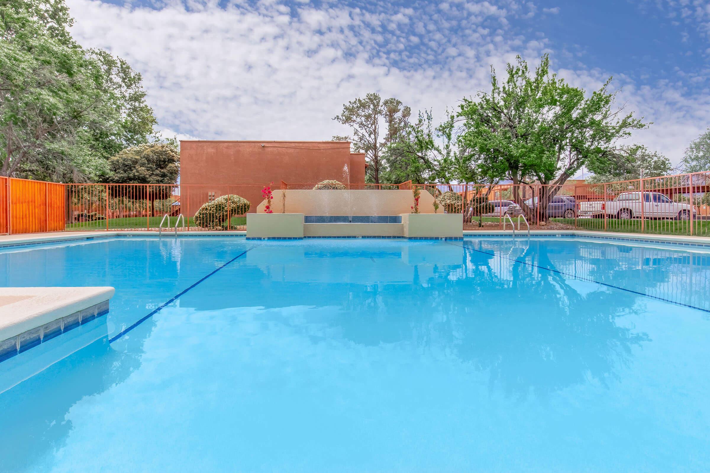 A clear blue swimming pool with a fountain feature in the center, surrounded by green trees and vibrant orange fencing. The sky above is partly cloudy, adding a serene atmosphere to the outdoor setting. Poolside ladders are visible, inviting for a refreshing swim.