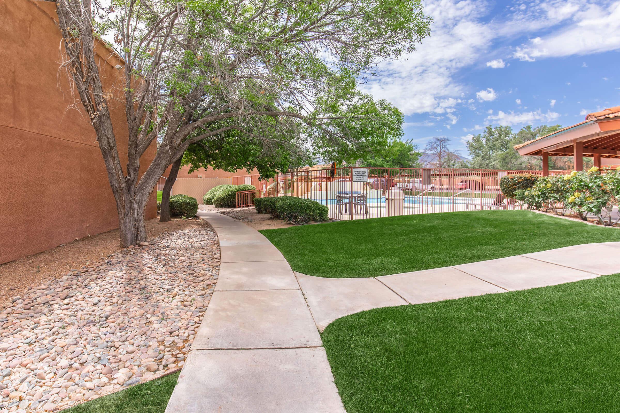 A well-maintained landscape featuring a stone path winding through lush green grass, bordered by a tree on one side. In the background, a fenced pool area is visible, surrounded by trees and shrubs, under a clear blue sky with scattered clouds.