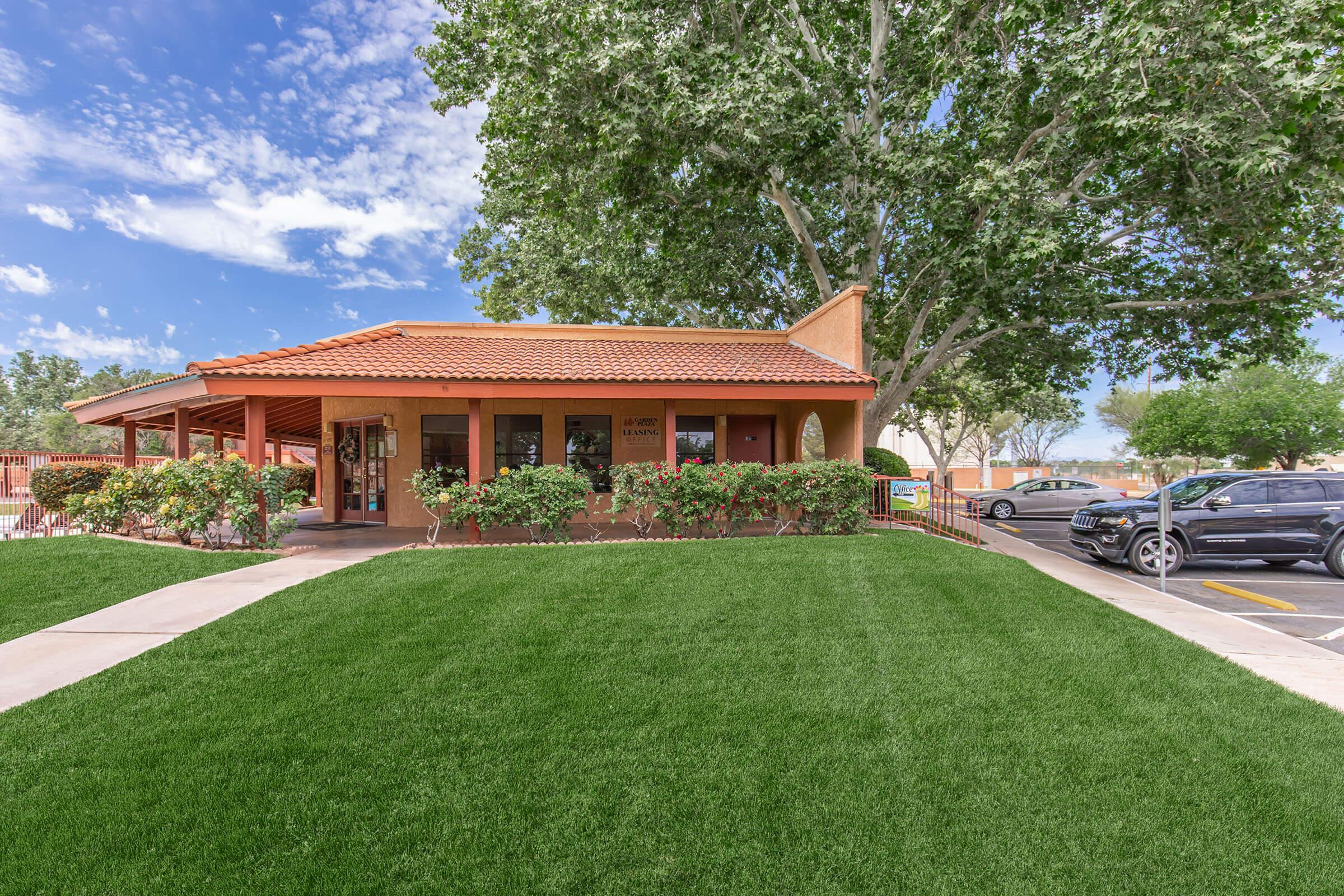 A single-story building with a red tile roof and large windows, surrounded by a well-maintained green lawn and shrubs. In the background, there are trees and a blue sky with scattered clouds. A parking area is visible to the right with vehicles parked.