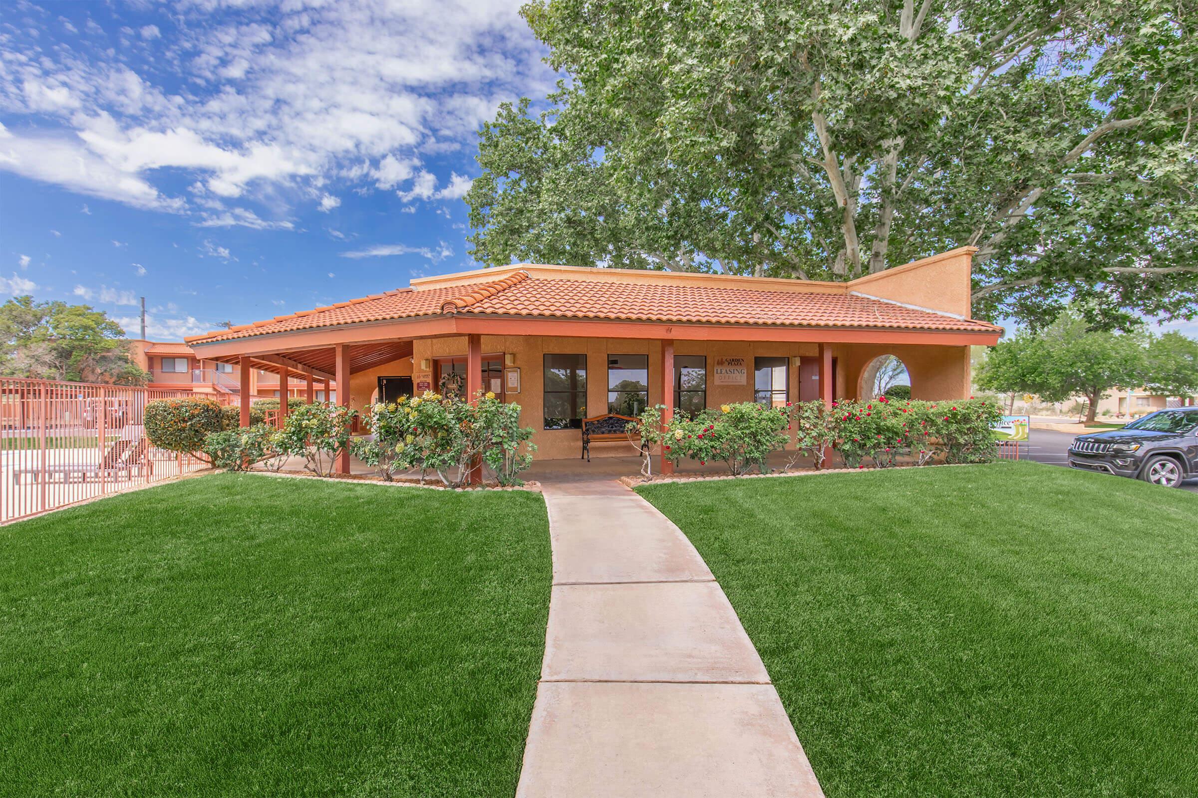 A single-story building with a terracotta roof, surrounded by lush green grass and flowering shrubs. A paved walkway leads to the entrance, and there are large windows allowing natural light. In the background, trees and a blue sky with scattered clouds can be seen.