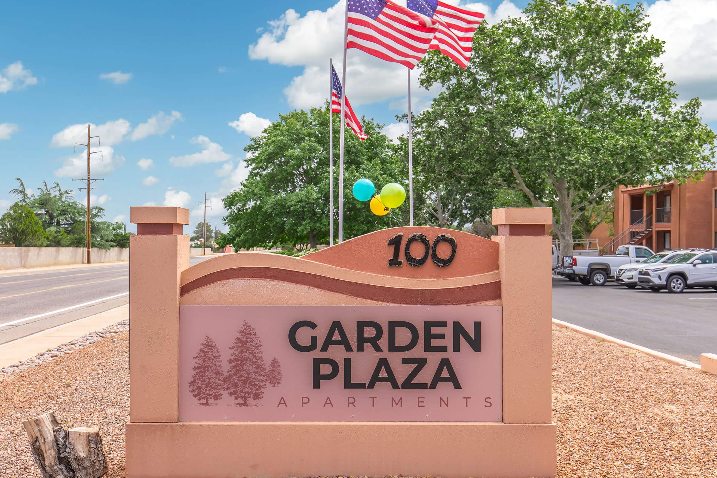 Sign for Garden Plaza Apartments at 100, featuring a welcoming design with trees and colorful balloons. Two American flags are displayed on tall poles beside the sign. The surrounding area includes a paved road, greenery, and parked vehicles, creating a vibrant community atmosphere.