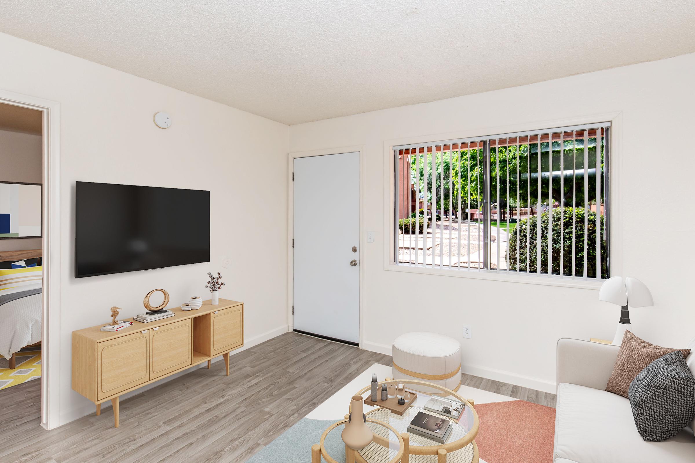 A cozy living room featuring a modern TV on a wooden stand, a light-colored sofa with decorative pillows, and a round coffee table with magazines. A large window allows natural light and provides a view of greenery outside. The walls are neutral, creating a bright and inviting atmosphere.