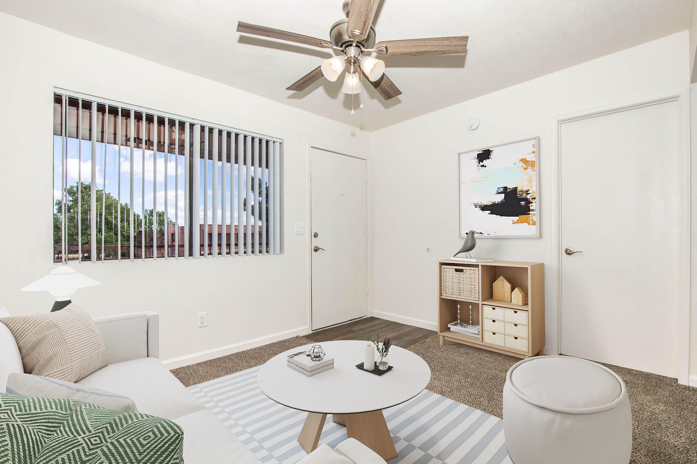 A modern living room with light-colored walls and carpet. There's a ceiling fan with wooden blades, a window with vertical blinds, and a door. A light-colored sofa is on the left, with a patterned throw pillow. A round coffee table is in the center, topped with decor items, and a small storage unit is against the wall.