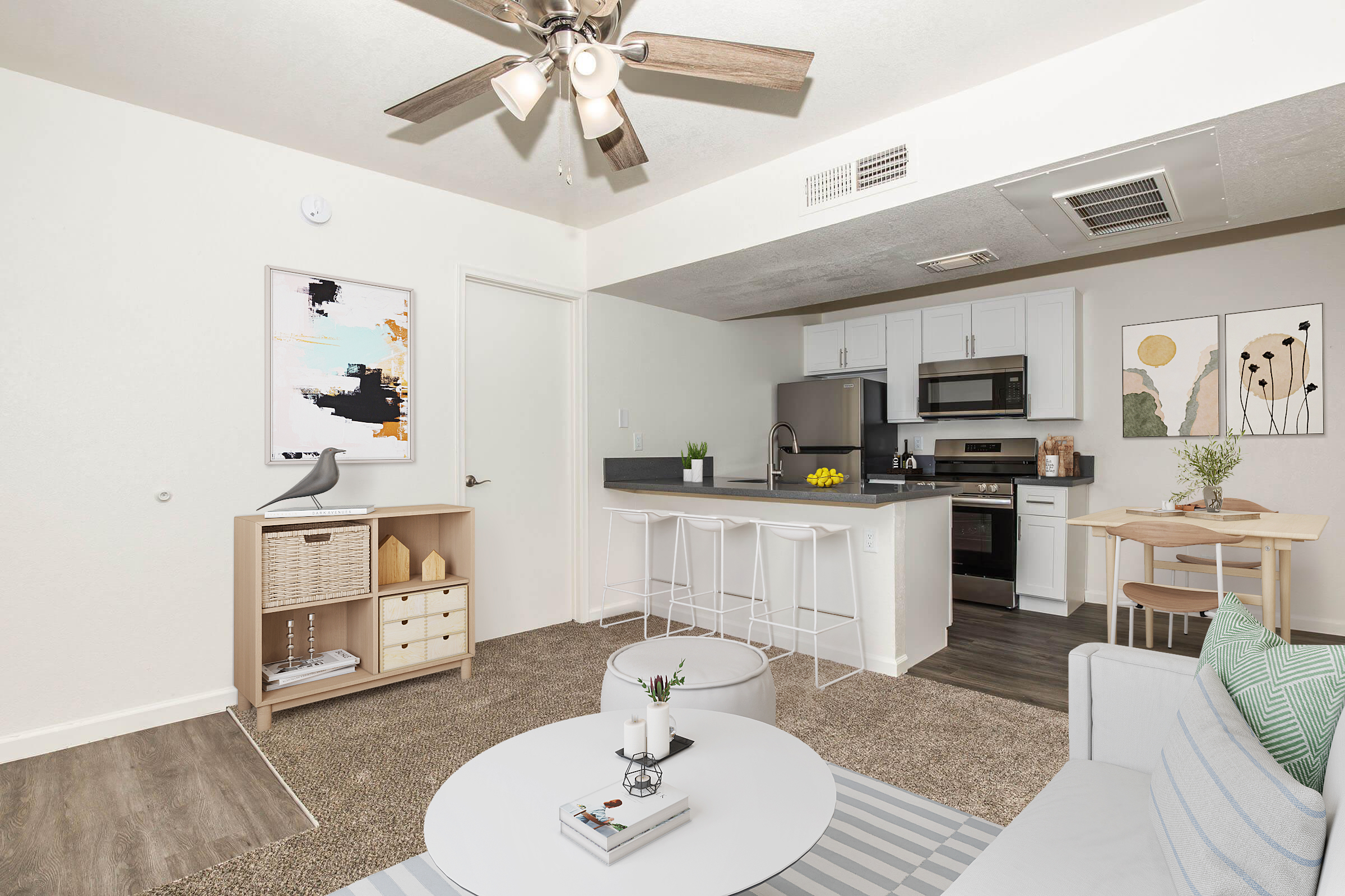 A modern, cozy living space featuring a light-colored couch, a round coffee table, and a stylish area rug. The kitchen is visible with white cabinetry and stainless steel appliances. There are decorative wall art pieces and a dining table set for two, adding a warm, inviting atmosphere.