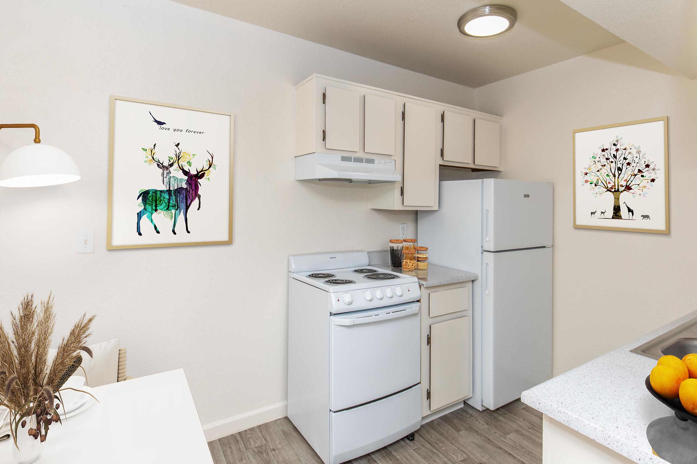 A modern, well-lit kitchen featuring a white stove and refrigerator, light-colored cabinets, and decorative wall art depicting colorful deer and a tree. The countertop is clutter-free, and a dining table with decorative elements is visible, enhancing the cozy atmosphere of the space.