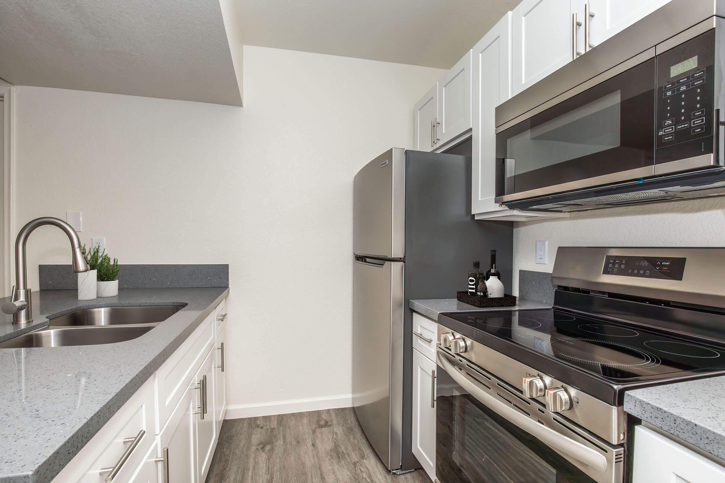 Modern kitchen featuring stainless steel appliances, including a refrigerator, microwave, and oven. The cabinetry is white with a sleek countertop and a double sink. A small plant adds a touch of greenery. The flooring is a light wood finish, contributing to a clean and contemporary look.