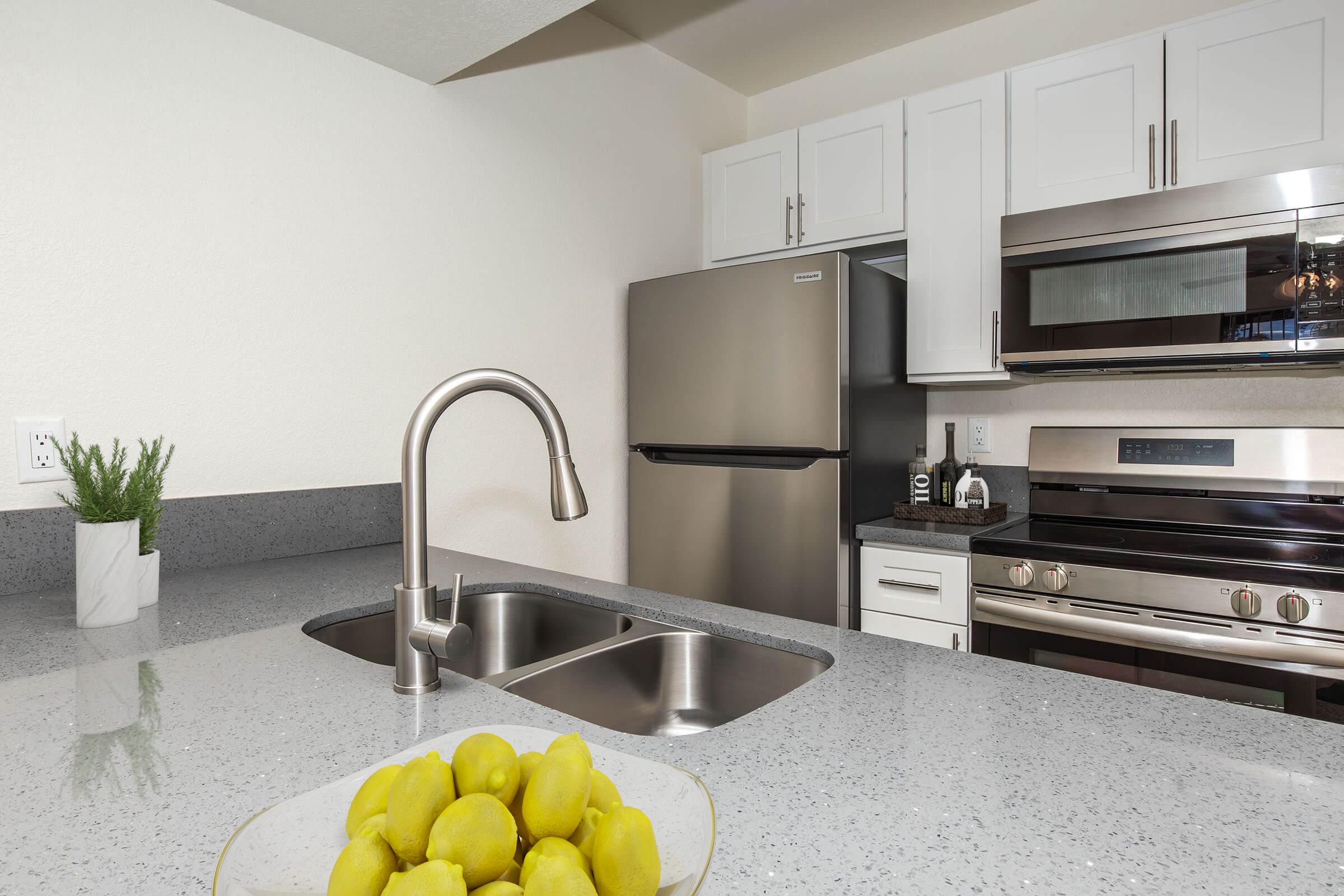 A modern kitchen featuring a stainless steel refrigerator and oven, white cabinetry, and a gray granite countertop. In the foreground, a glass bowl holds bright yellow lemons, and a small potted plant adds a touch of greenery. The space is well-lit and designed for contemporary living.