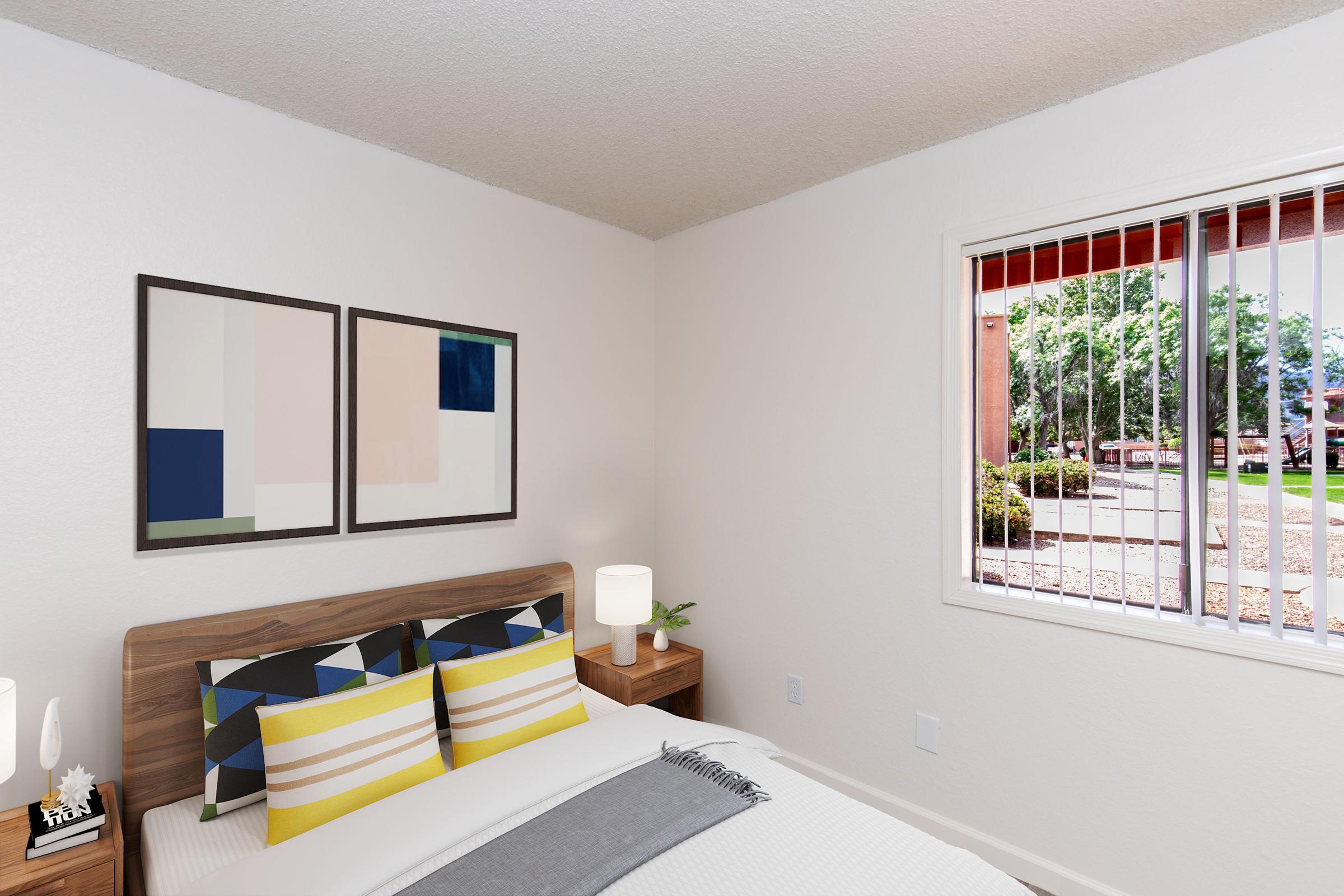 A cozy bedroom featuring a bed with patterned pillows, two framed artworks on the wall, a bedside table with a lamp and decorative items, and a window with blinds displaying a view of a landscaped exterior. The room has a neutral color palette and modern decor elements.