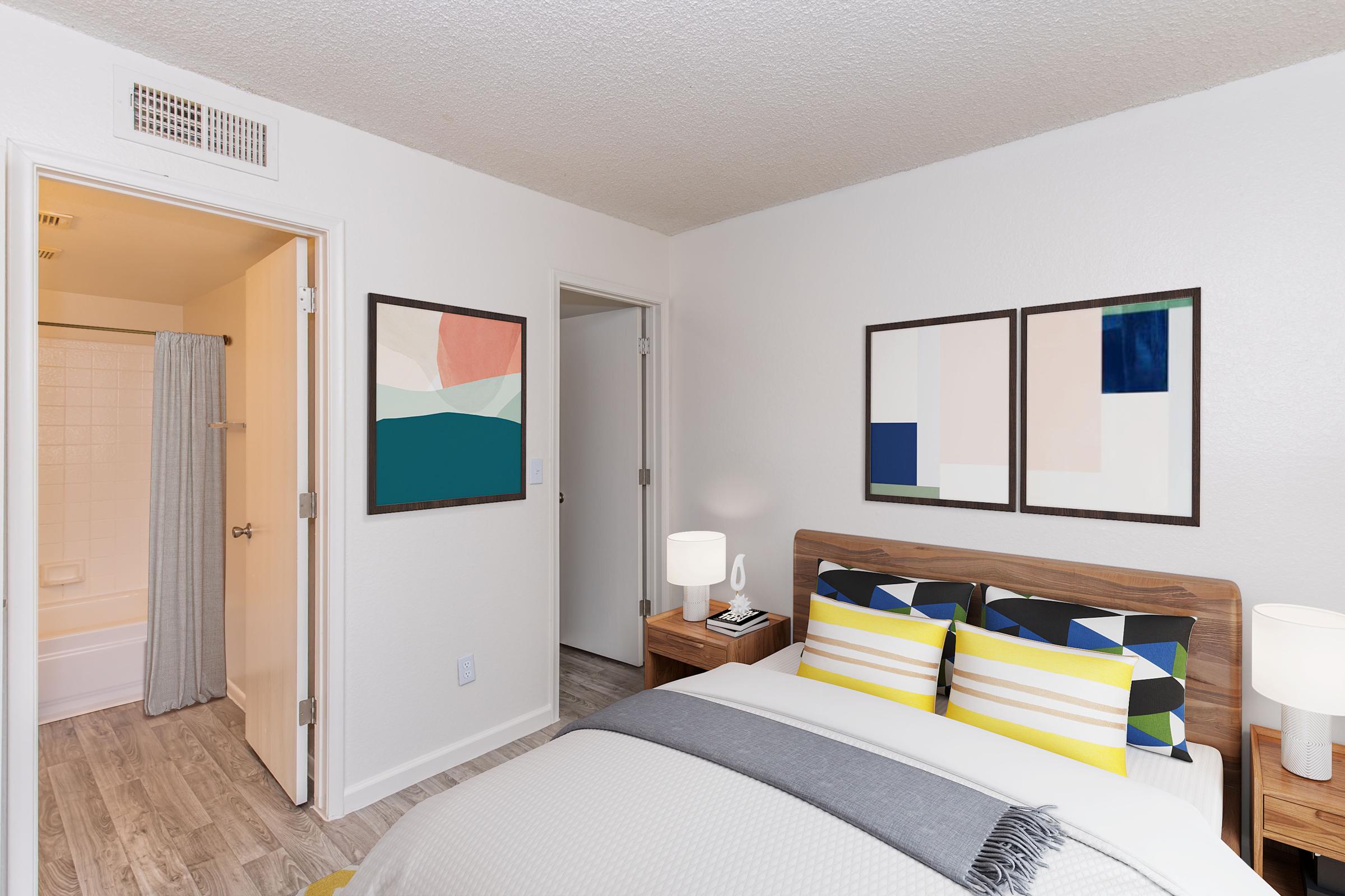 A modern bedroom featuring a bed with colorful striped pillows, a stylish gray throw, and two contemporary lamps on nightstands. The walls are decorated with abstract art pieces. A bathroom is visible through an open door, and there is a closet door on the opposite side, showcasing a clean and inviting space.