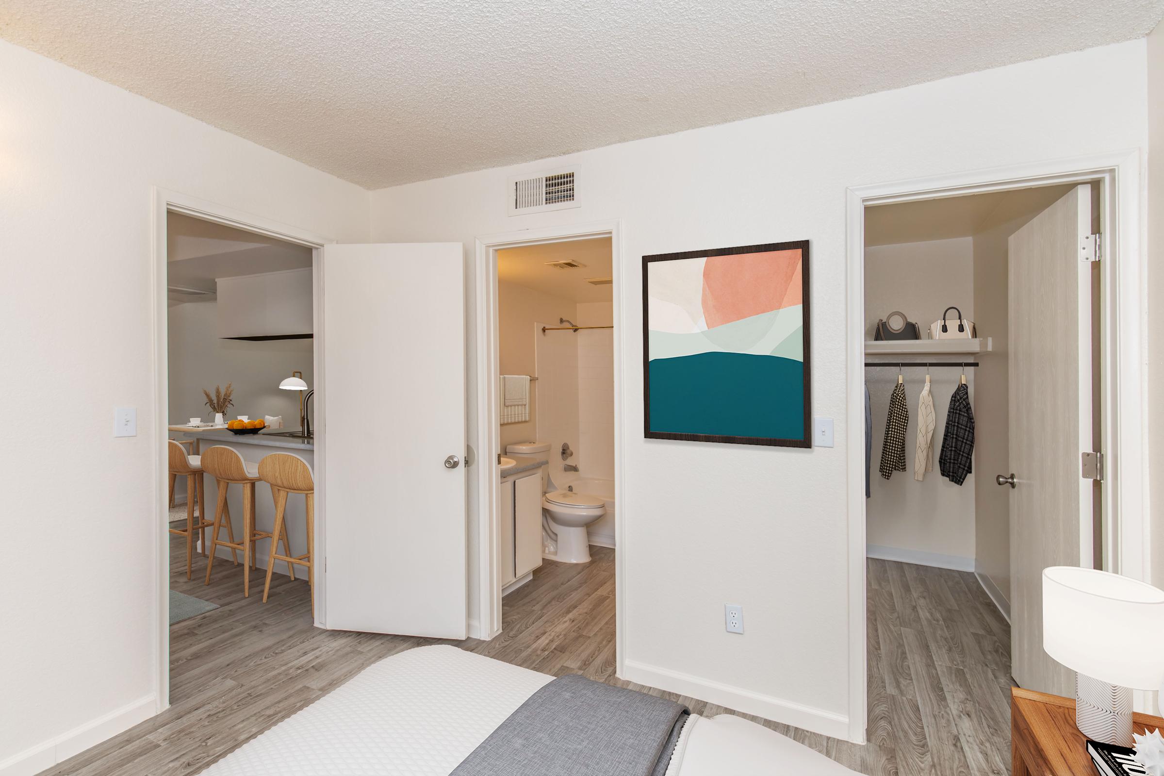 A cozy bedroom featuring a simple bed with gray bedding, a framed abstract artwork on the wall, and a door leading to a closet on the right. An open bathroom door can be seen in the background. A small table lamp sits on a nightstand, and the overall decor is modern and minimalistic.