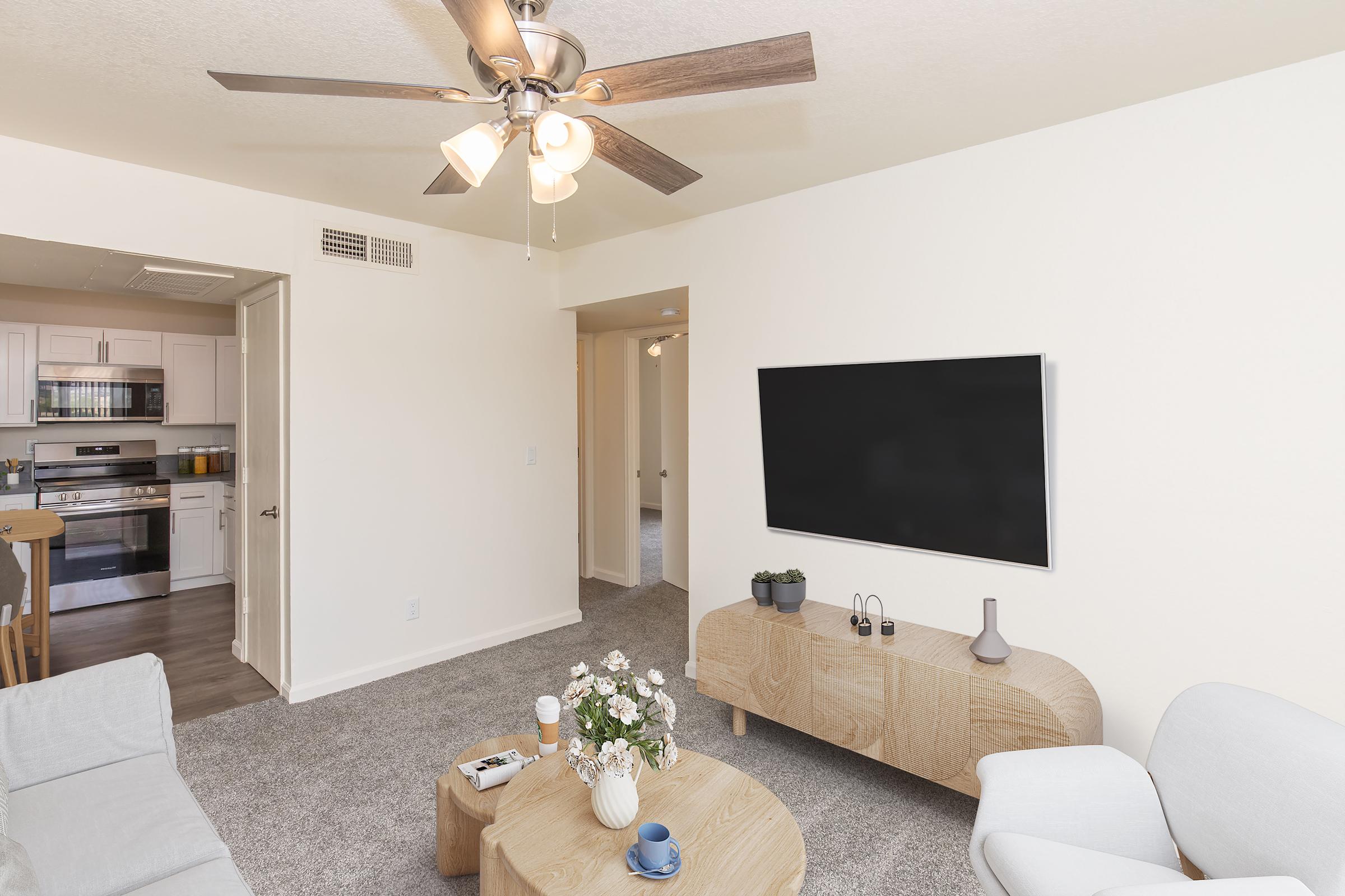A bright and modern living room with light-colored walls and carpet. It features a ceiling fan, a large flat-screen TV mounted on the wall, a wooden coffee table with a vase of flowers, and a comfortable seating area with a light gray sofa and armchair. A glimpse of a kitchen is visible in the background.