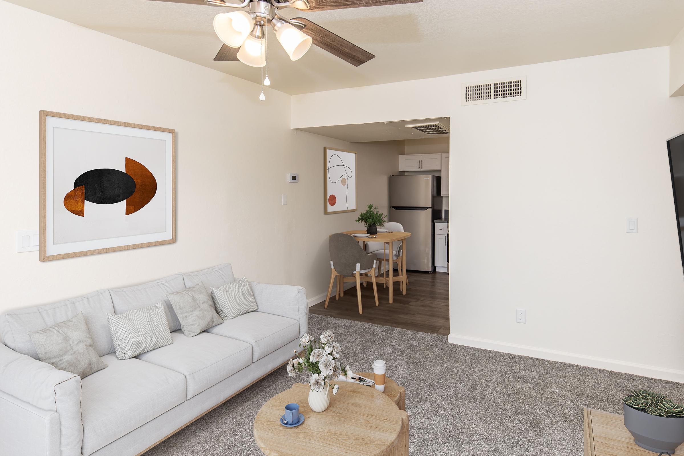A bright, modern living space featuring a light gray sofa with decorative pillows, a circular wooden coffee table, and a plant. An abstract artwork hangs on the wall. In the background, a small dining area with a round table and four chairs is visible, along with a kitchen space. The flooring is light-colored and the walls are painted a soft beige.